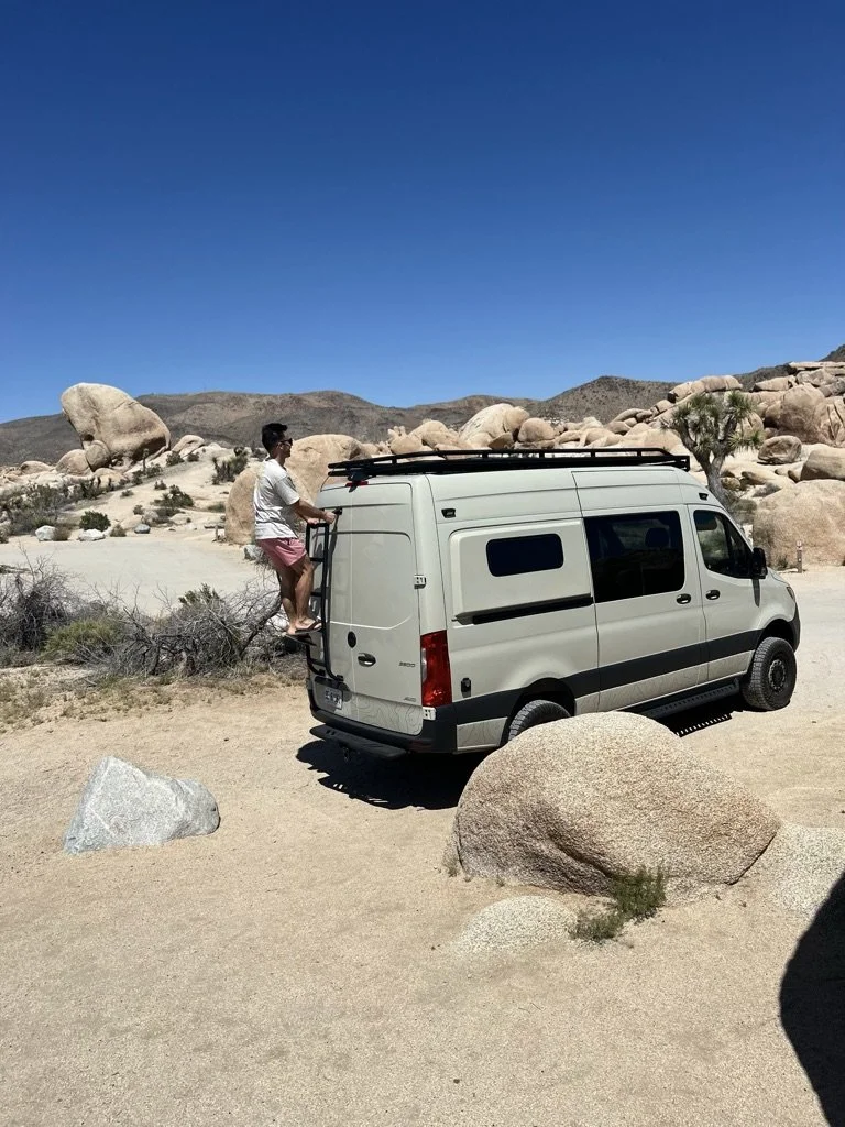 Person climbing a ladder on a camper van in a desert landscape with rocks and clear blue sky.