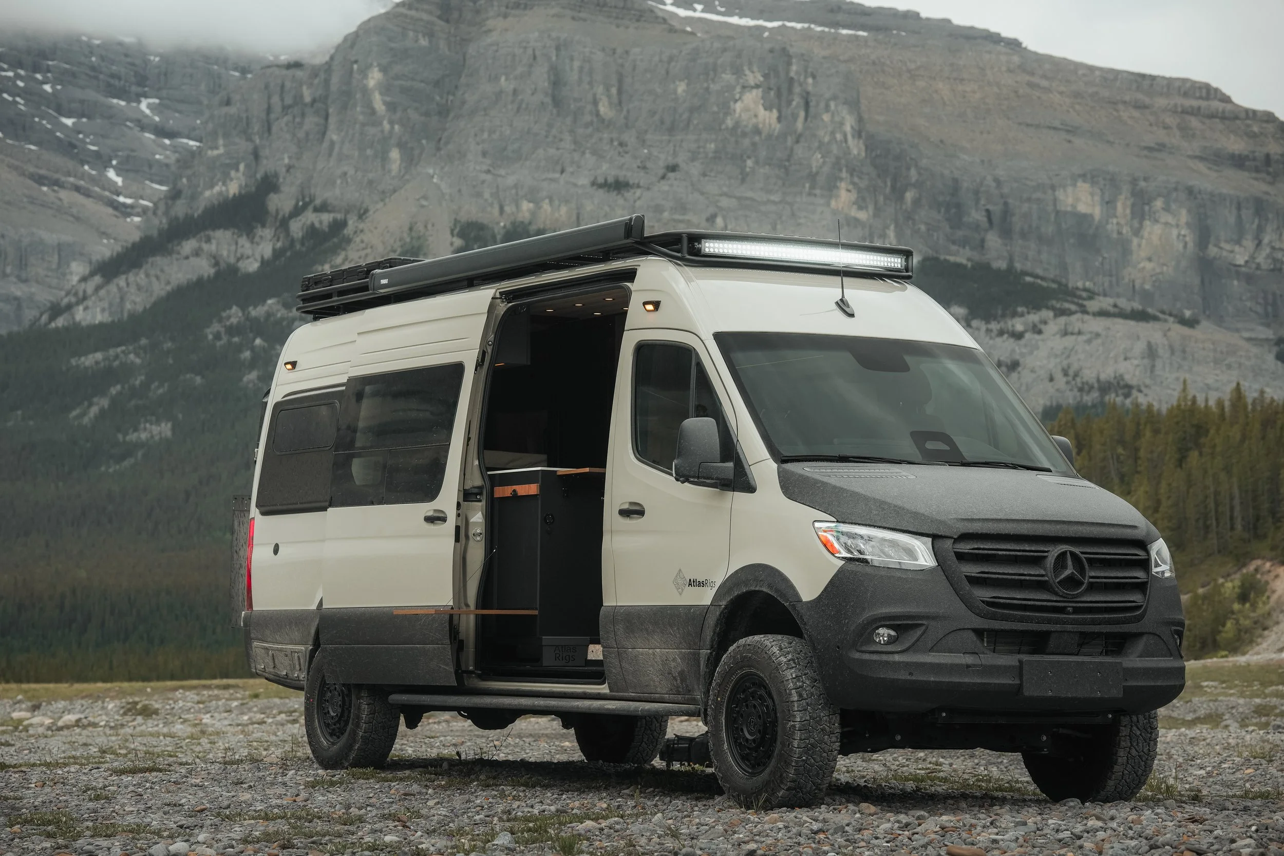 A white camper van with black tires and a black front grille parked on a gravel surface with a mountain range in the background.