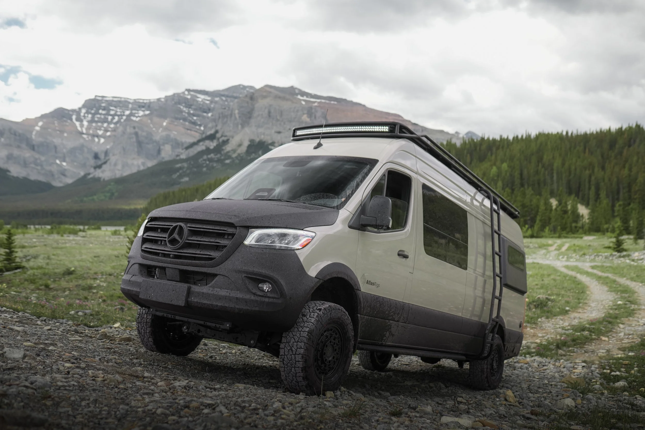 A white Mercedes-Benz van with off-road tires parked on rocky terrain in front of a mountain landscape with green trees and snow patches on the peaks, under cloudy skies.