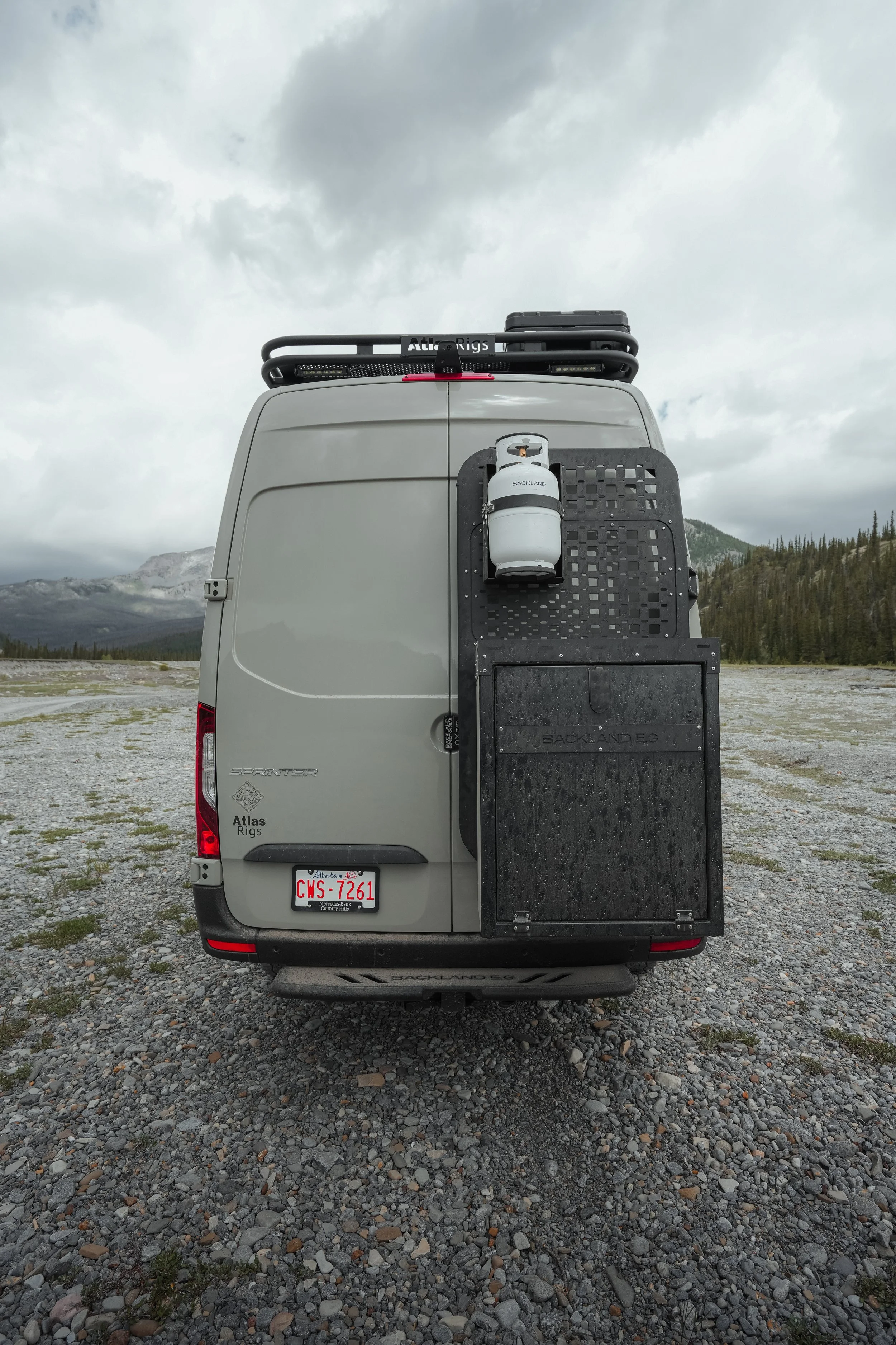 The image shows the back of a gray van with a propane tank mounted on a black rack attached to the rear door. The van is parked on a gravel surface with mountains and trees in the background under a cloudy sky.