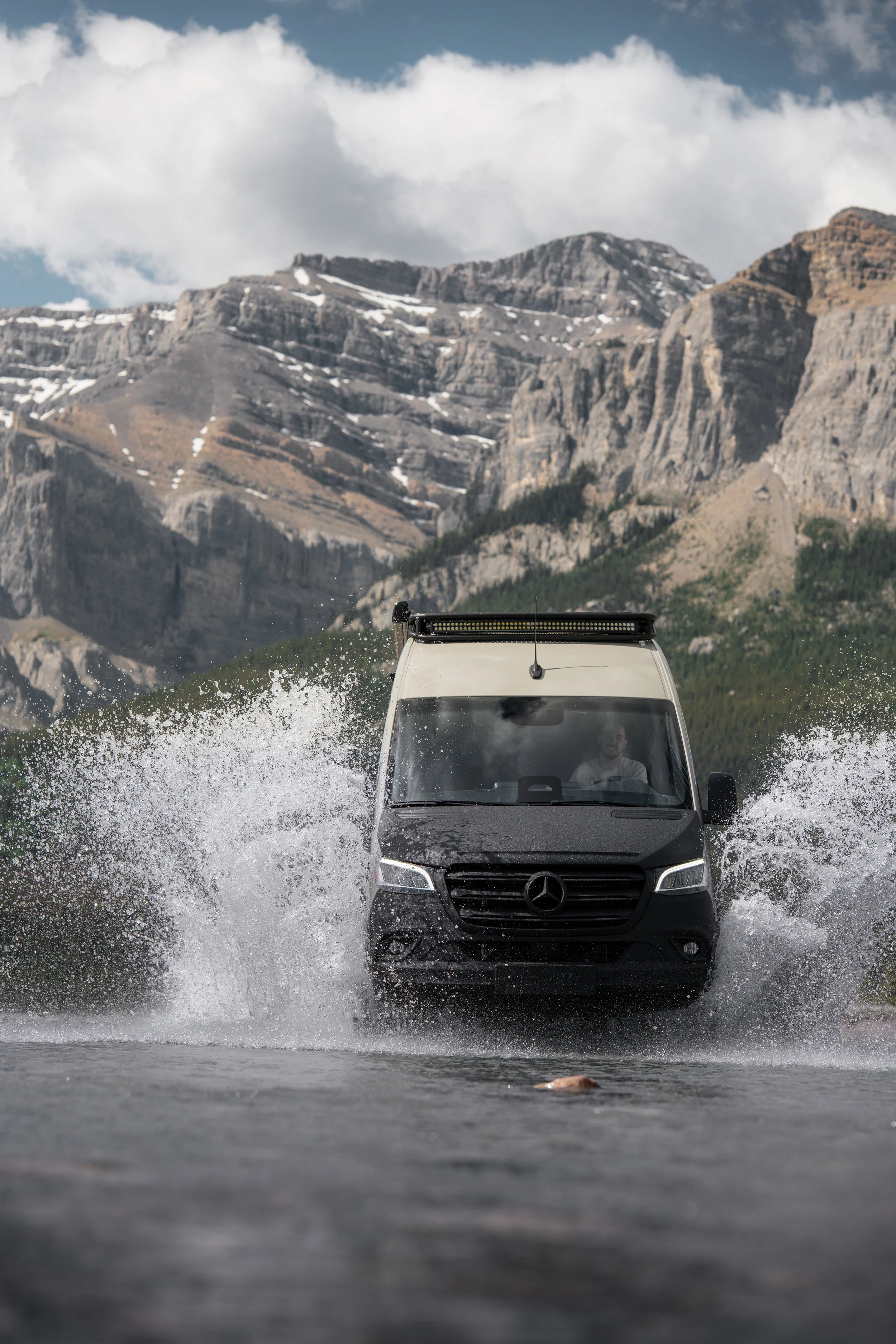 A black Mercedes-Benz van driving through a body of water, creating splashes, with mountains and clouds in the background.