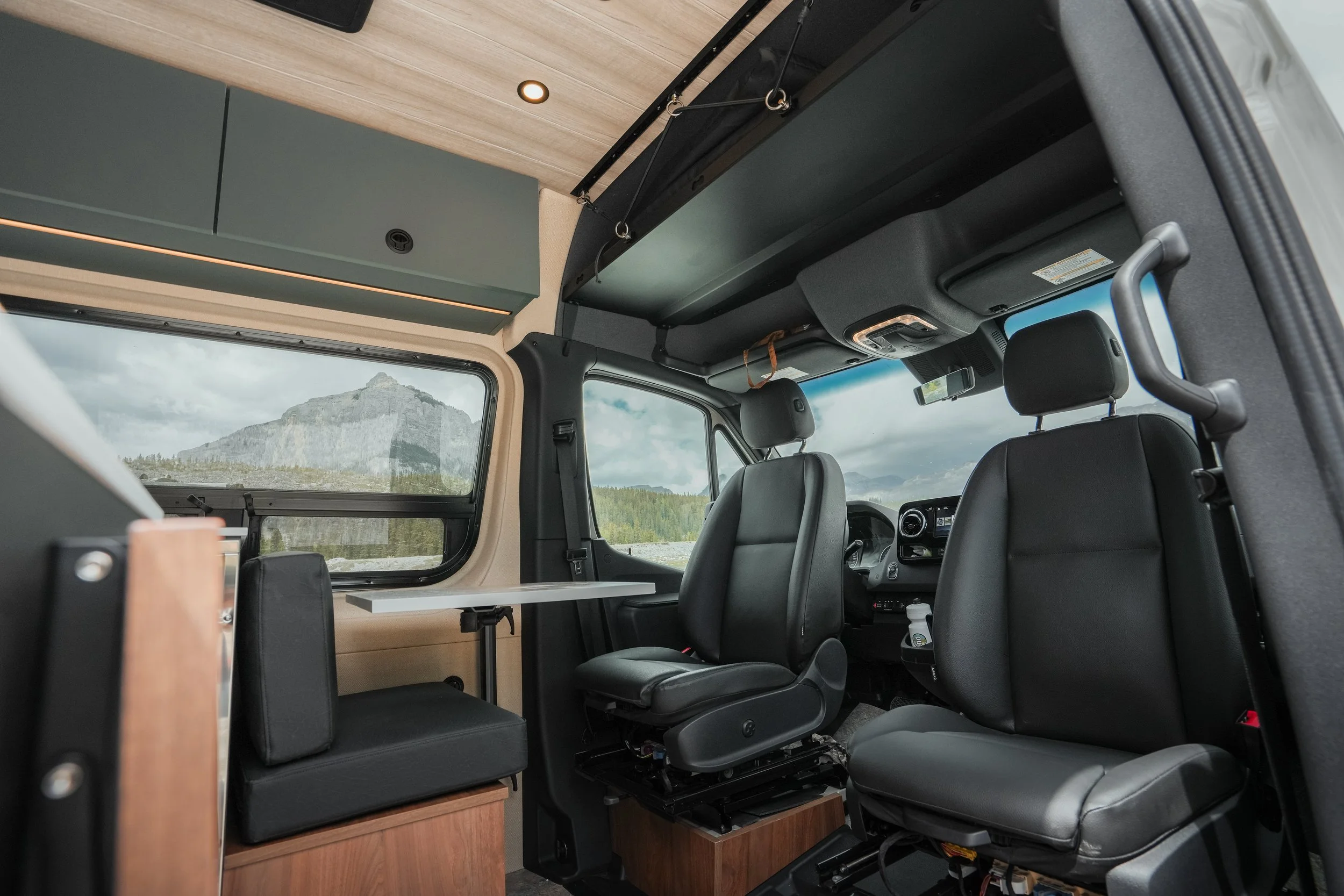Inside a camper van, showing the driver's seat and a passenger seat. Overhead storage cabinets, a small table, and a scenic mountain view through the window.
