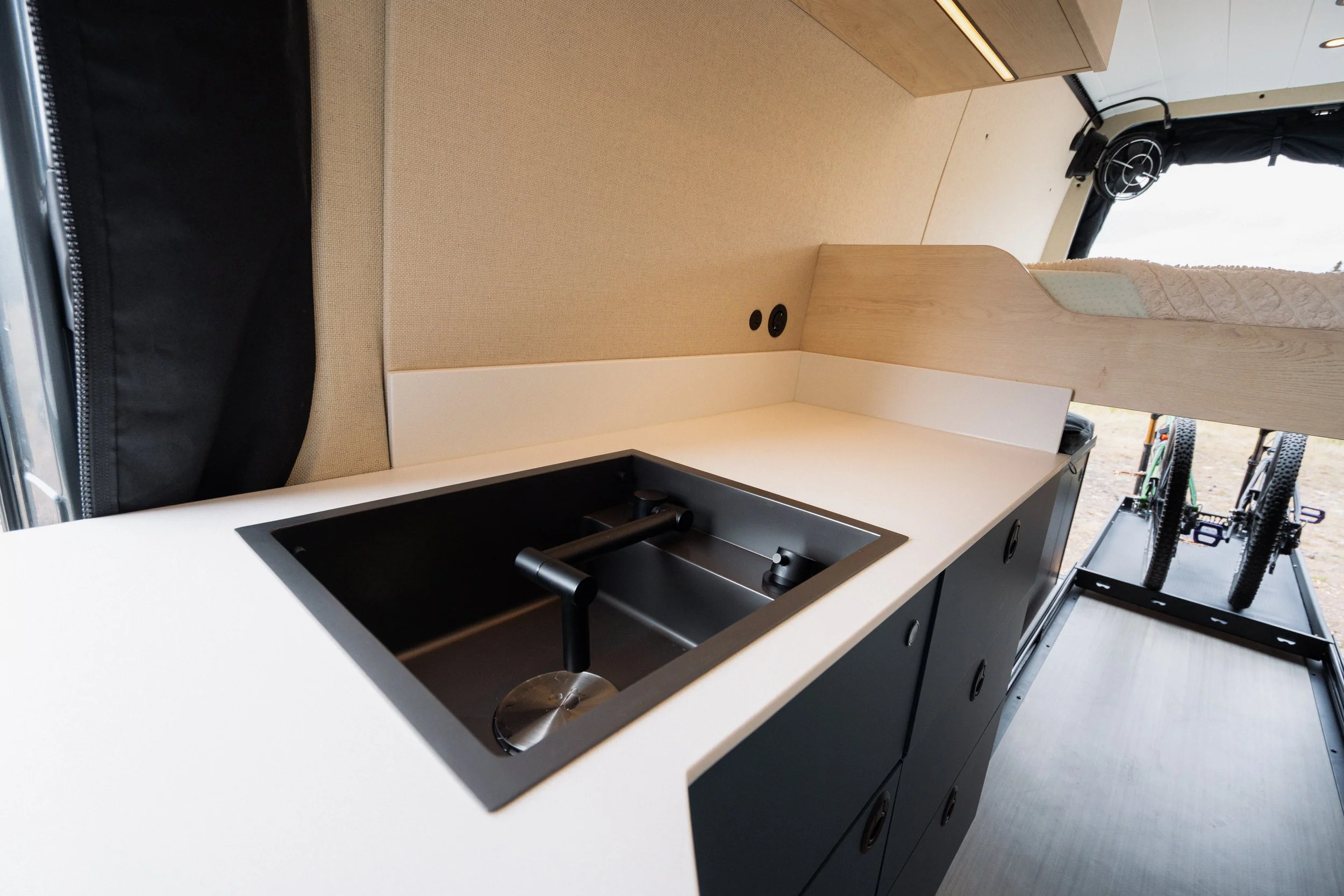 Interior of a compact kitchen in a camper van with a black sink, beige wall, wooden panel, and external view of bicycles outside.