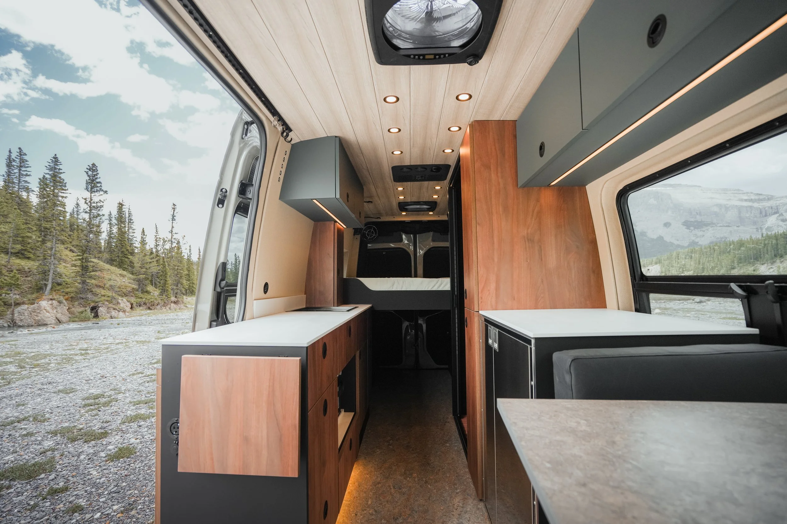 Interior view of a camper van with wood paneling, cabinets, countertops, and windows showing a natural landscape with trees and mountains outside.