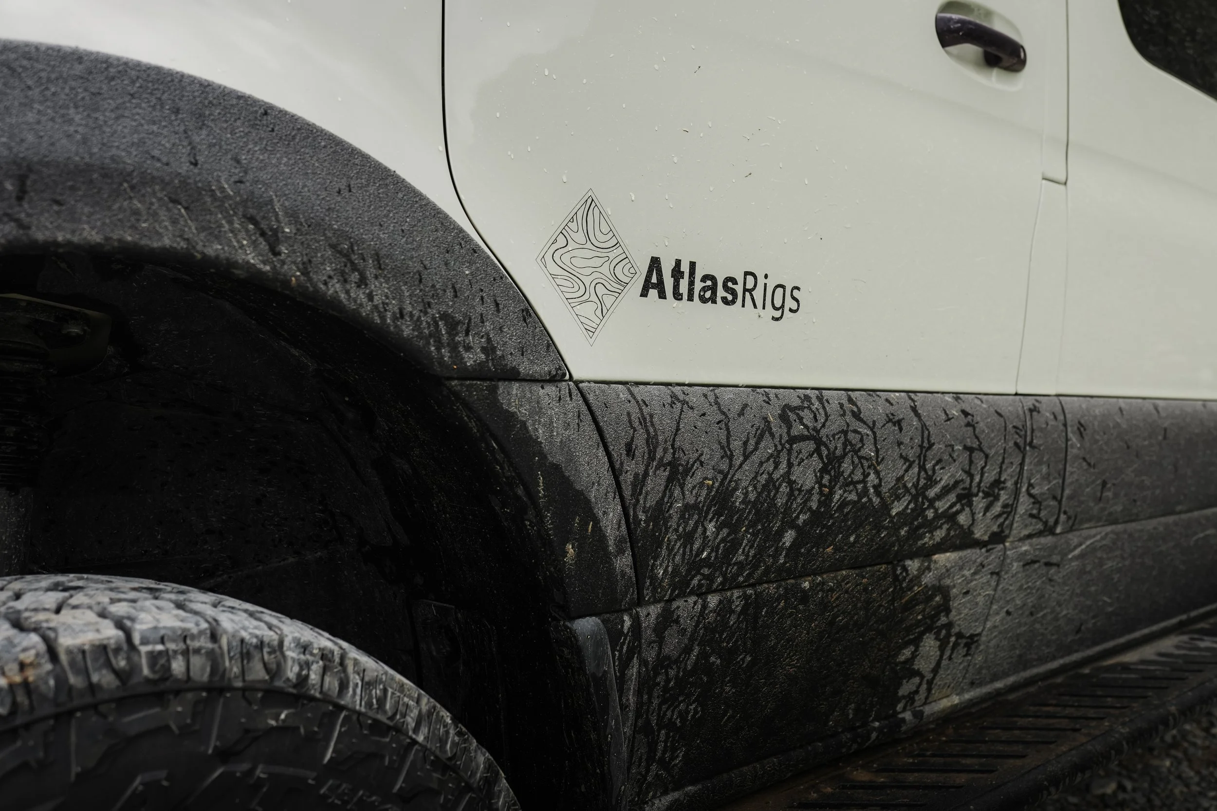 Close-up of a black tire and a white vehicle with a decal that reads 'AtlasRigs' on the side panel, with water droplets on the surface.
