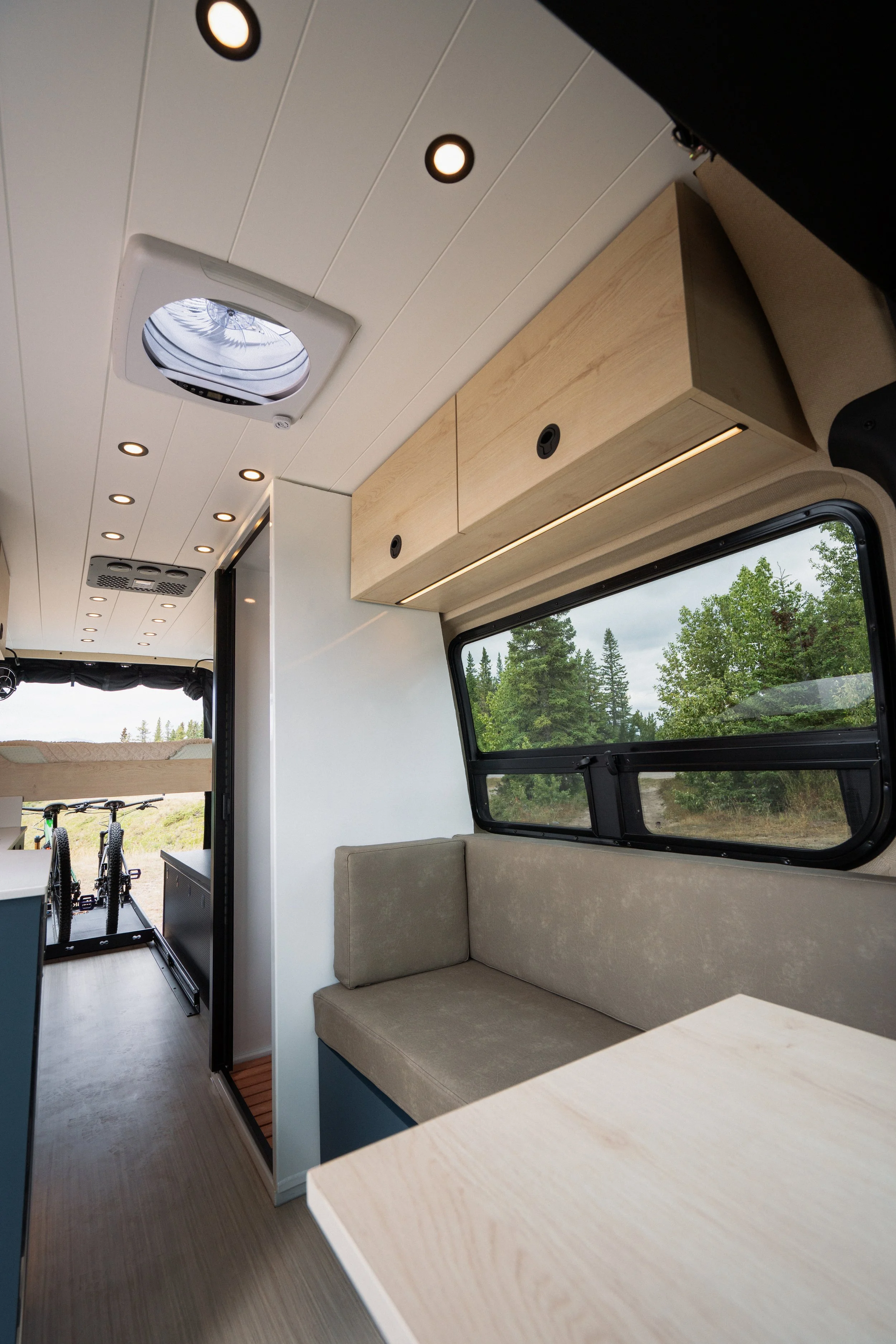 Interior of a camper van with a beige sofa, window with a view of trees, mounted wooden cabinets, ceiling lights, and a ceiling vent.