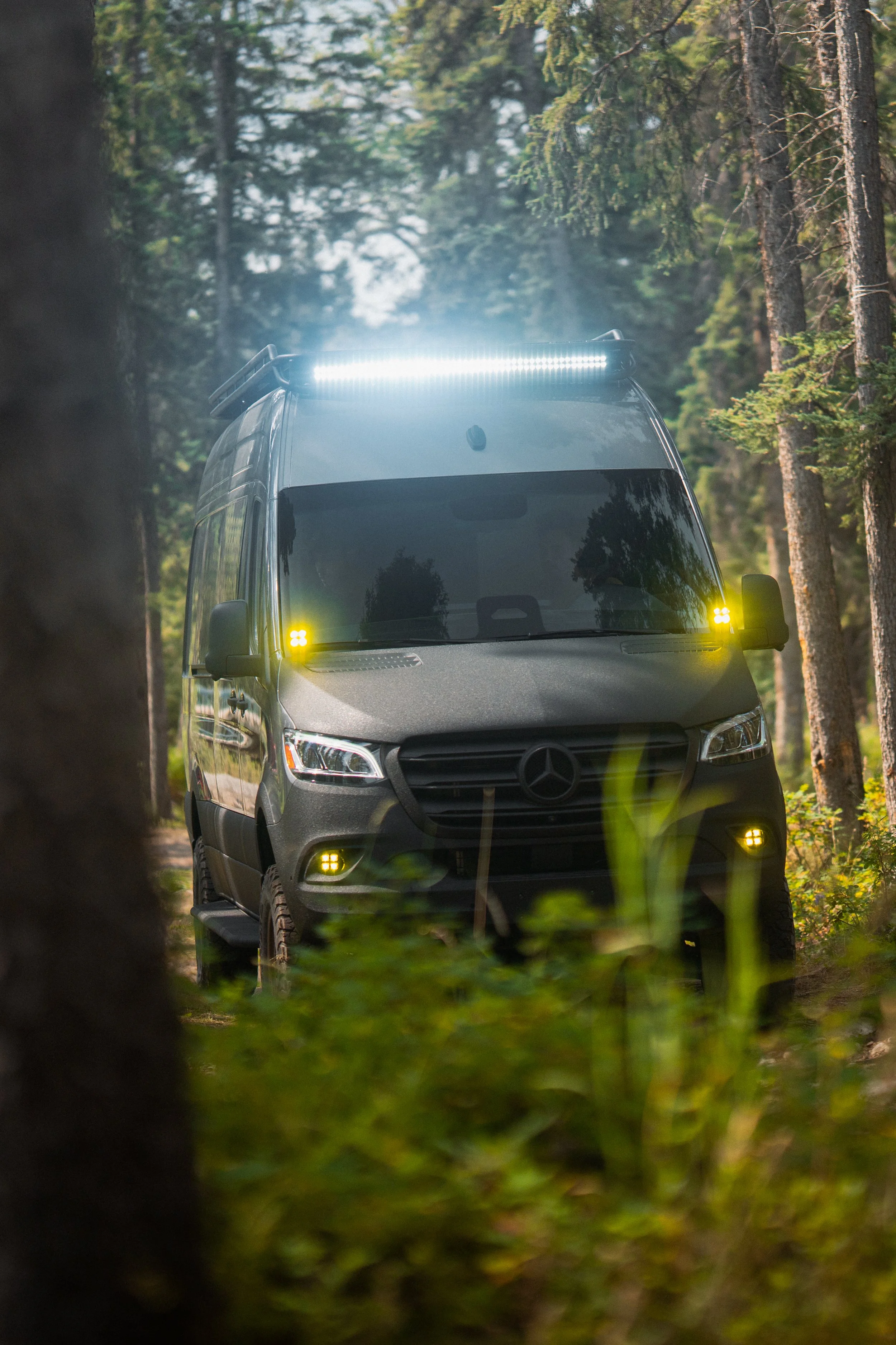 A gray Mercedes-Benz van with off-road tires and yellow and white emergency lights, driving through a dense forest with green trees and sunlight filtering through.