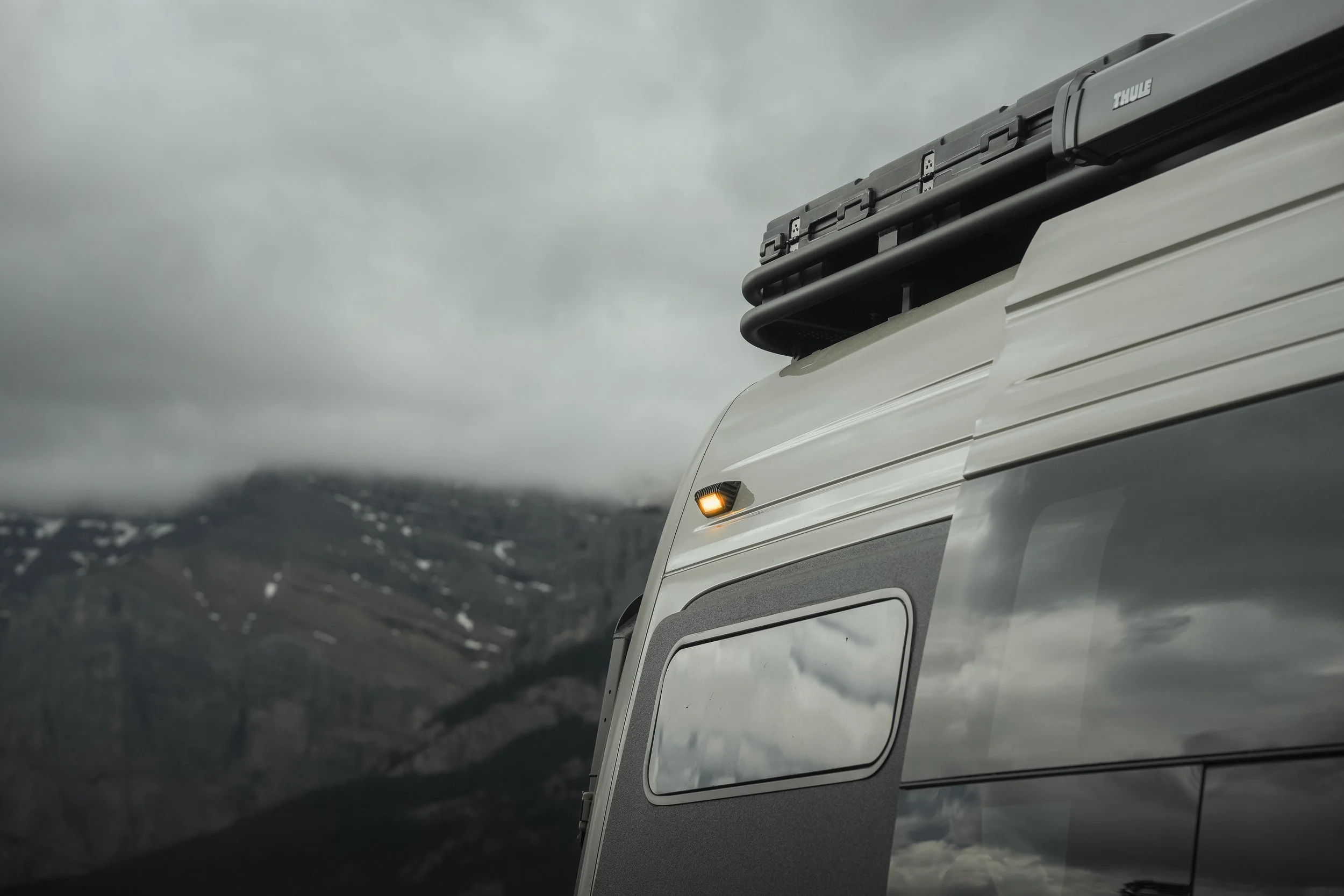 Front part of a white vehicle with roof storage and a small amber side marker light, against a backdrop of mountains and cloudy sky.