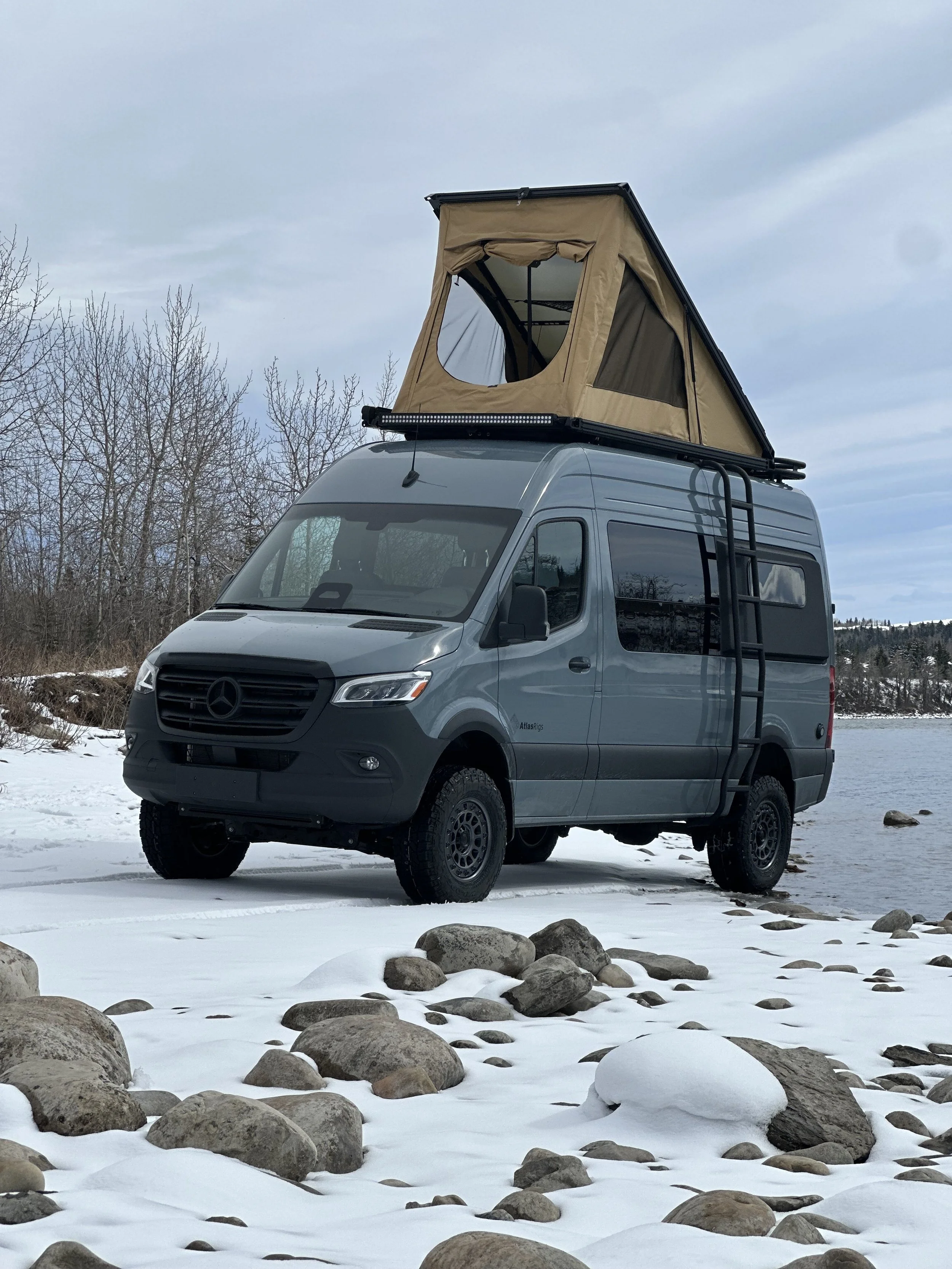 A gray camper van with a rooftop tent set up on a snowy landscape near a body of water, with leafless trees and a cloudy sky in the background.