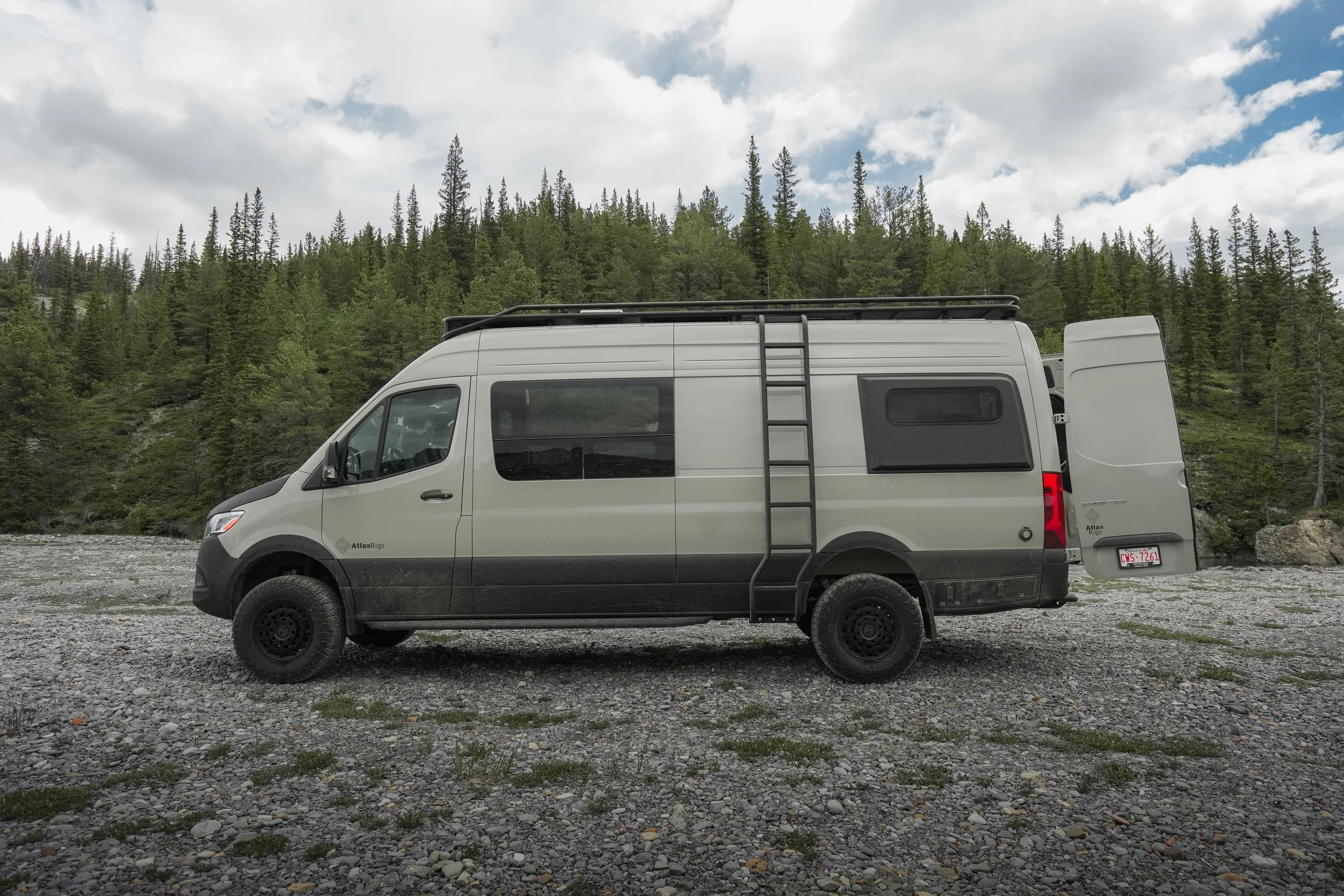 A white camper van parked on a gravel area near a forest with tall green trees and cloudy sky.