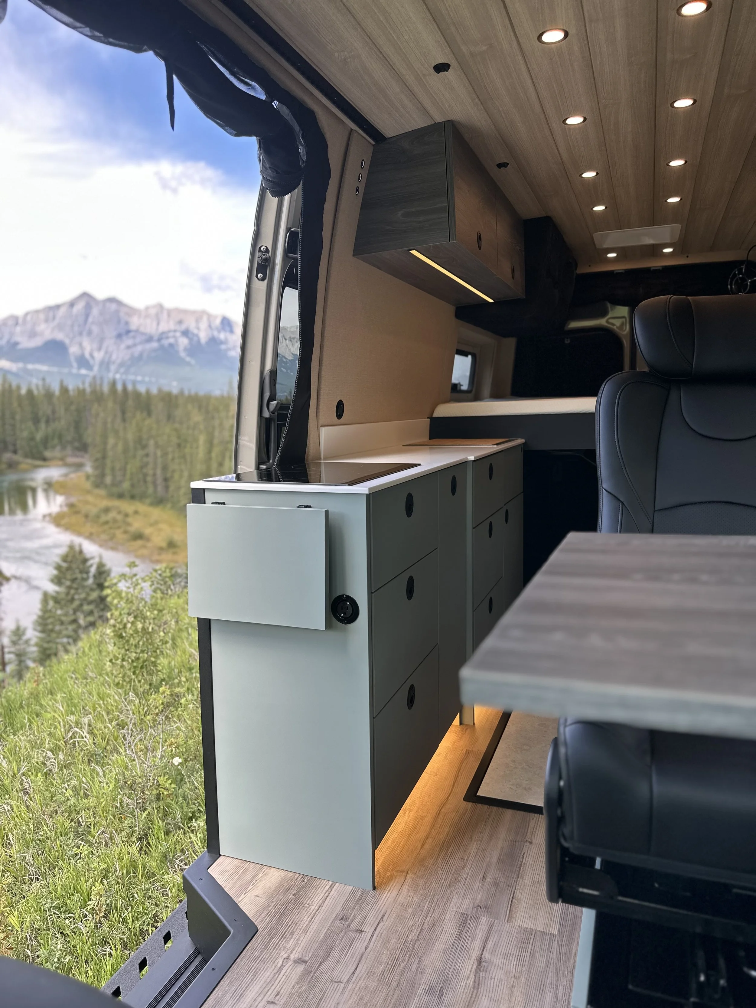 Interior of a modern camper van with a view of mountains and trees through the open window, featuring a kitchen area with cabinets, countertop, and seating with a black leather chair.