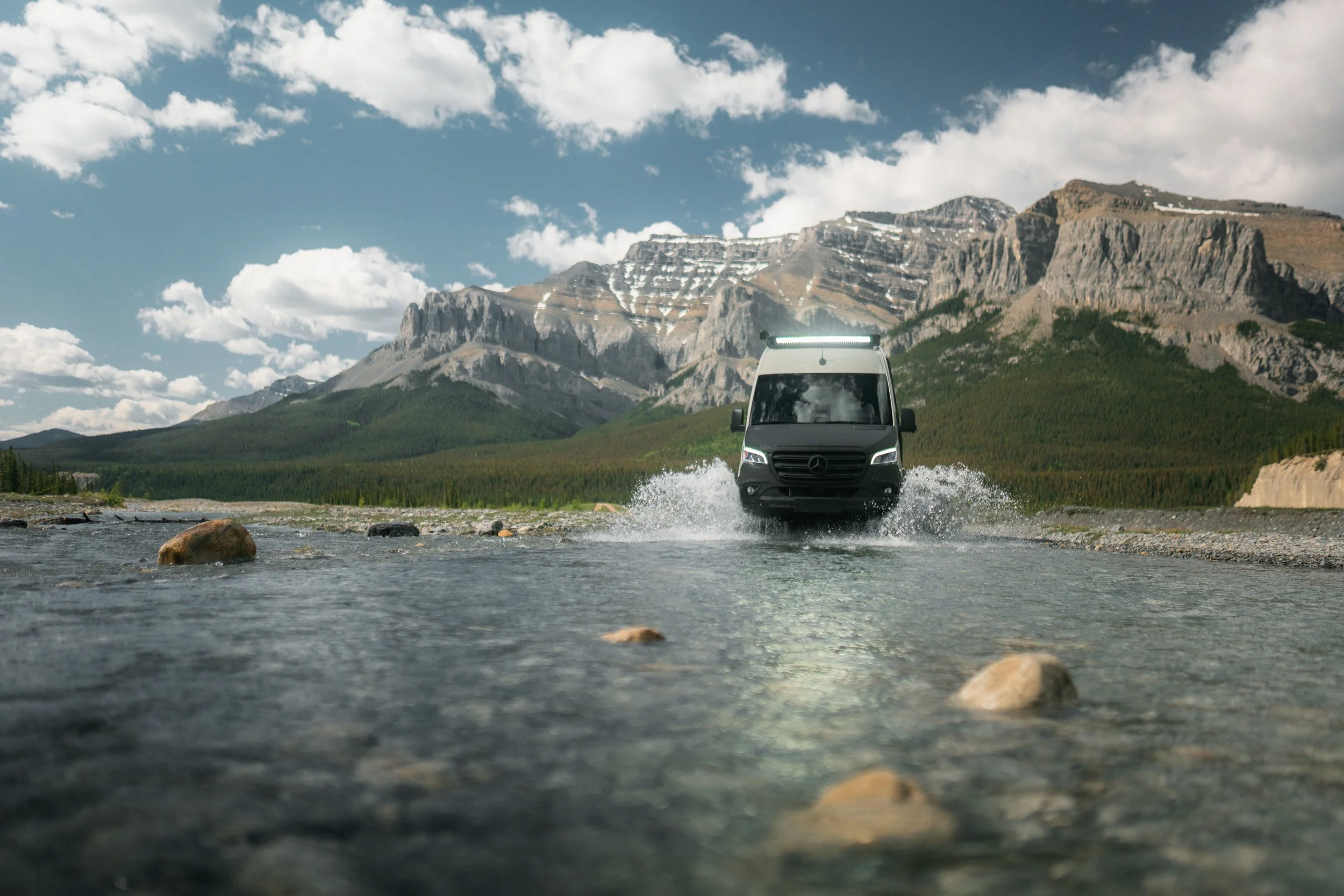 A black van crossing a shallow river with mountains and a partly cloudy sky in the background.
