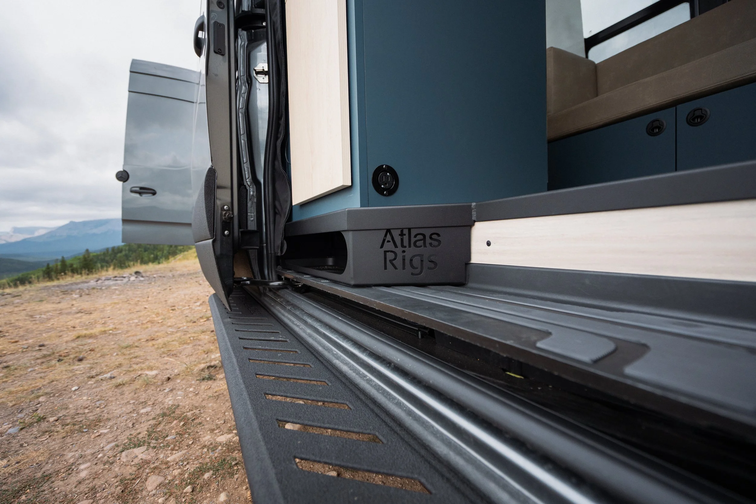 Close-up of a truck bed with a blue cabinet or storage unit labeled 'Atlas Rigs' and part of a seating area with brown cushions, set against an outdoor landscape with mountains and cloudy sky.