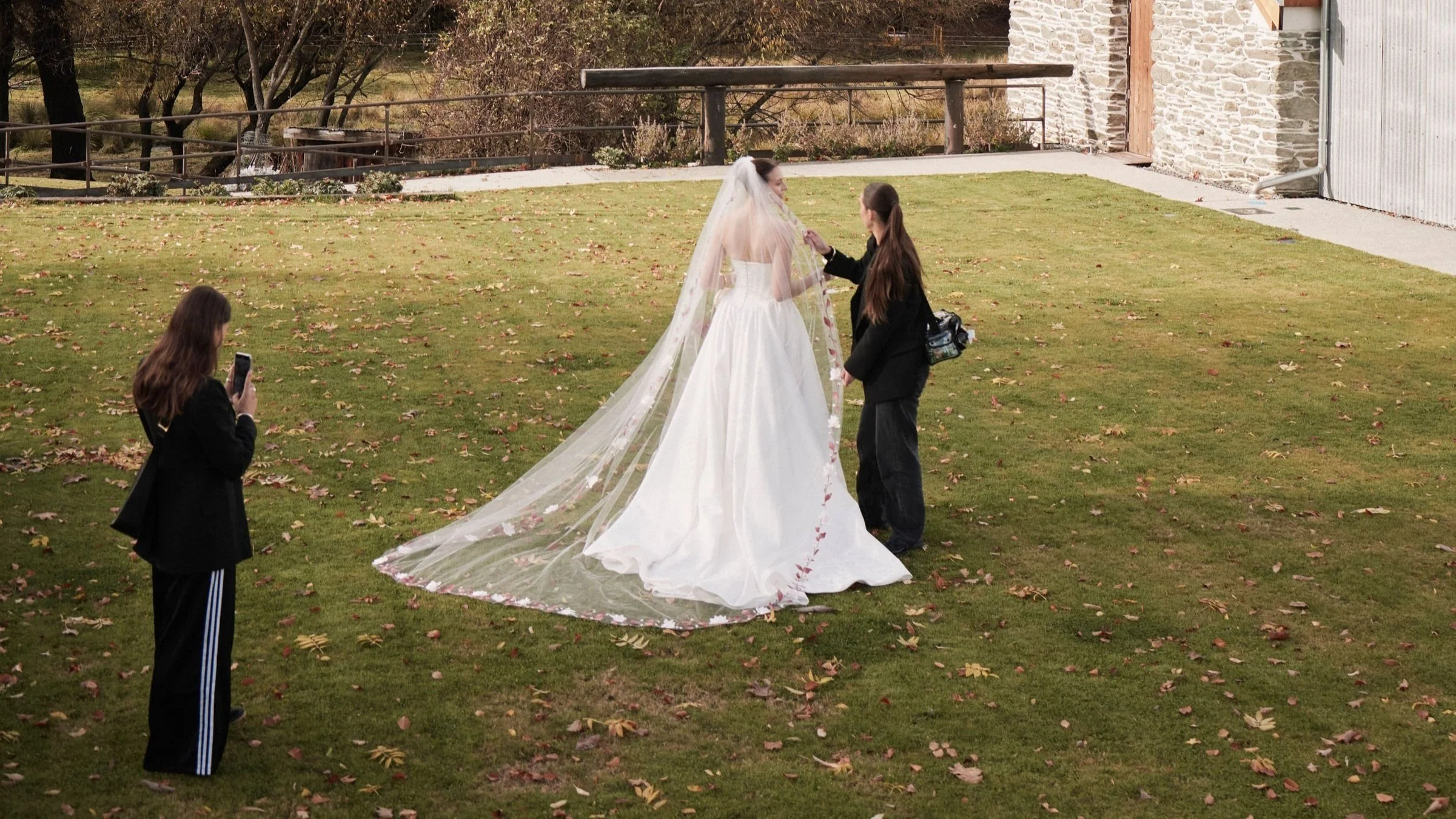 Behind-the-scenes wide shot at Merryfield, Arrowtown — Nicola filming on iPhone as makeup artist Liv King touches up a bride in Hera Couture during a styled shoot