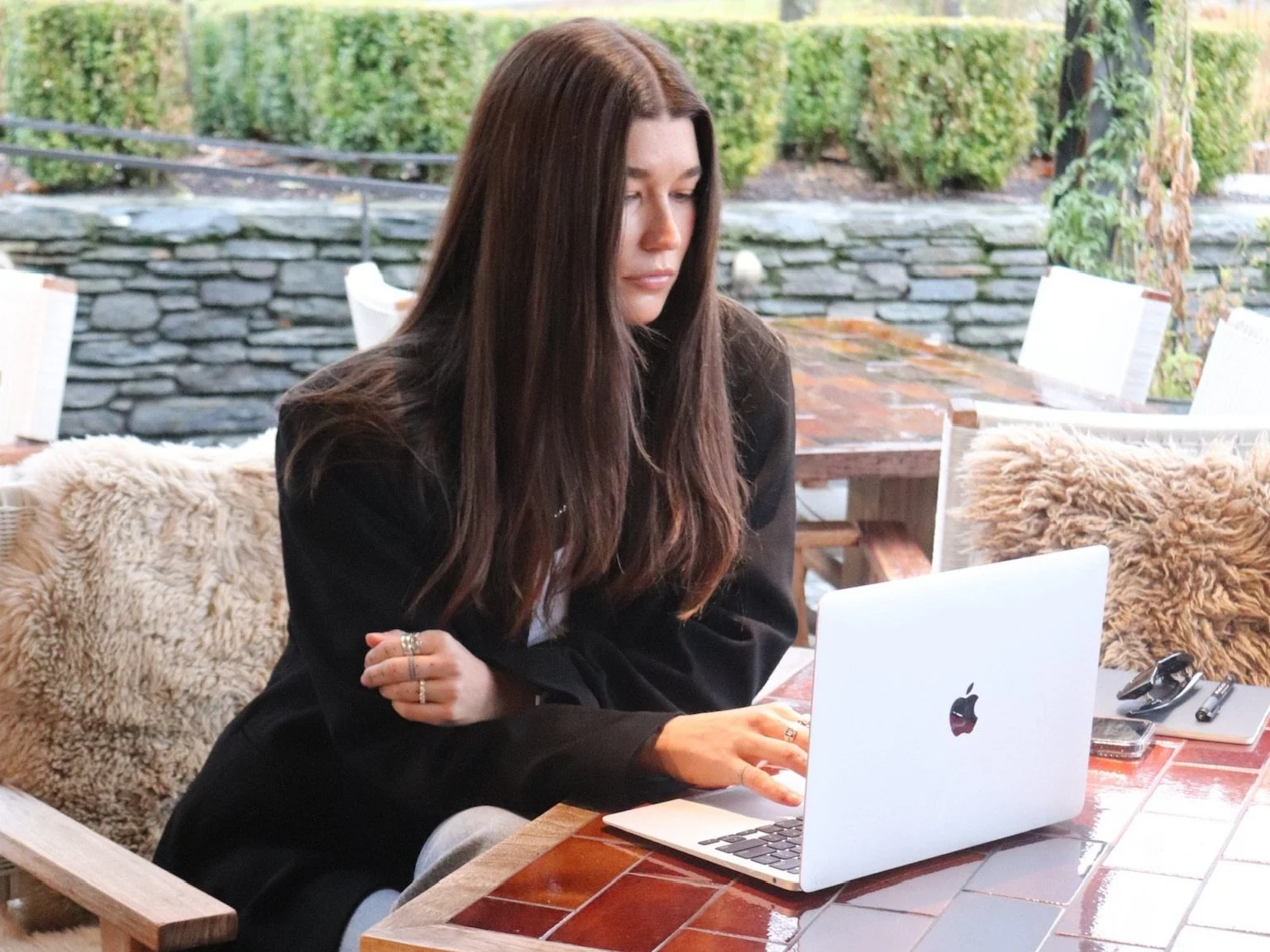 Nicola working at a table at Ayrburn - hair down, wearing a blazer and jeans, with MacBook open and notebook, sunglasses, and fur throws in the background.