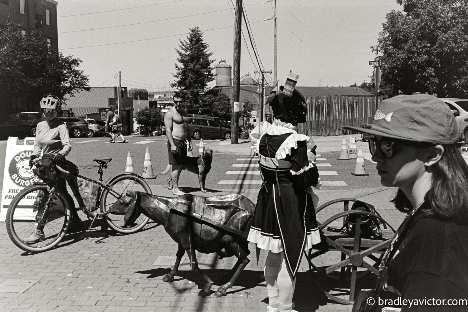 Photo by Bradley A. Victor. A candid urban tableau captured on black and white film. People fill the frame in the fore mid and background, gazing in different directions. One woman notices the camera and smiles, while a shirtless man stares at her.