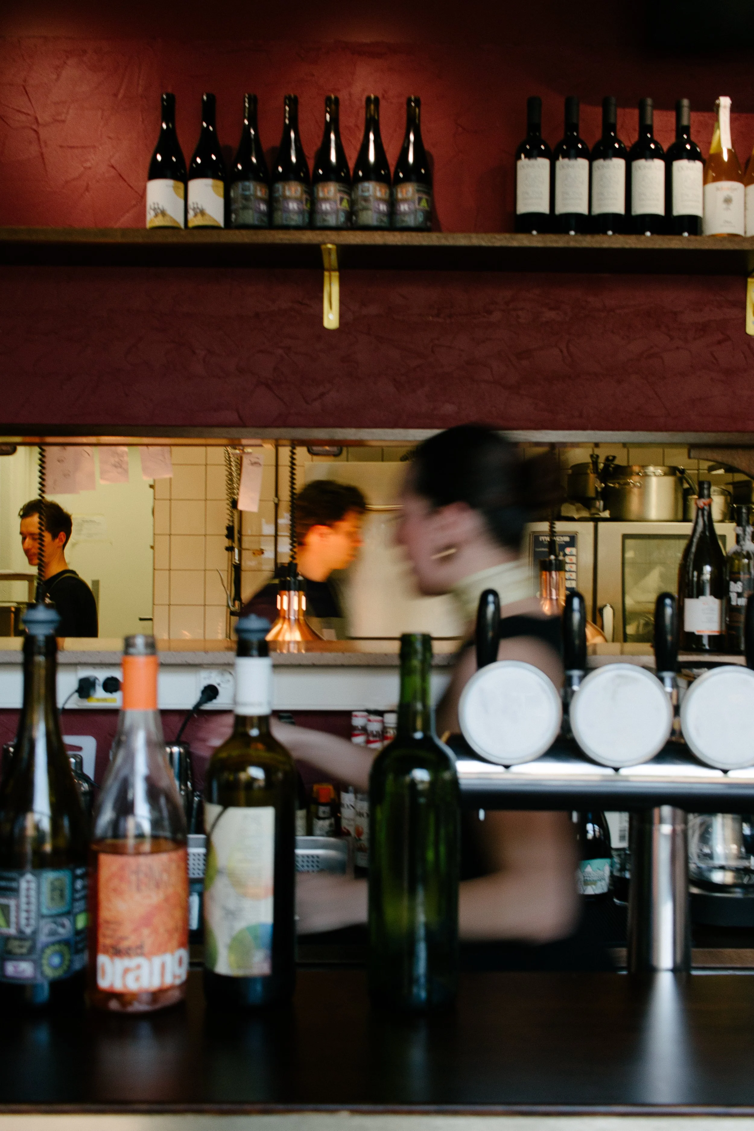 Bar with bottles of wine and liquor on shelves, bartender preparing drinks, kitchen staff working in the back, and blurred patrons at the bar.
