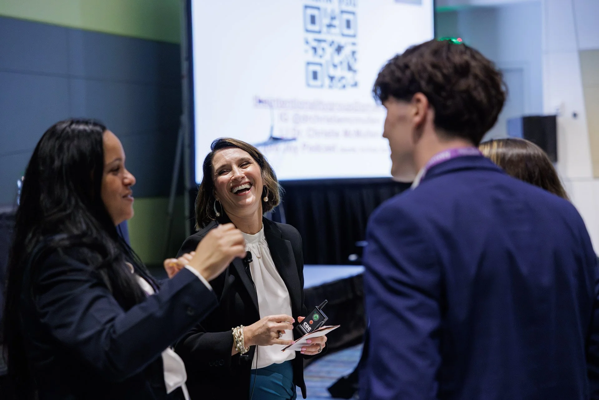 Group of four people engaged in conversation at conference or event, with a QR code displayed on a screen in the background.