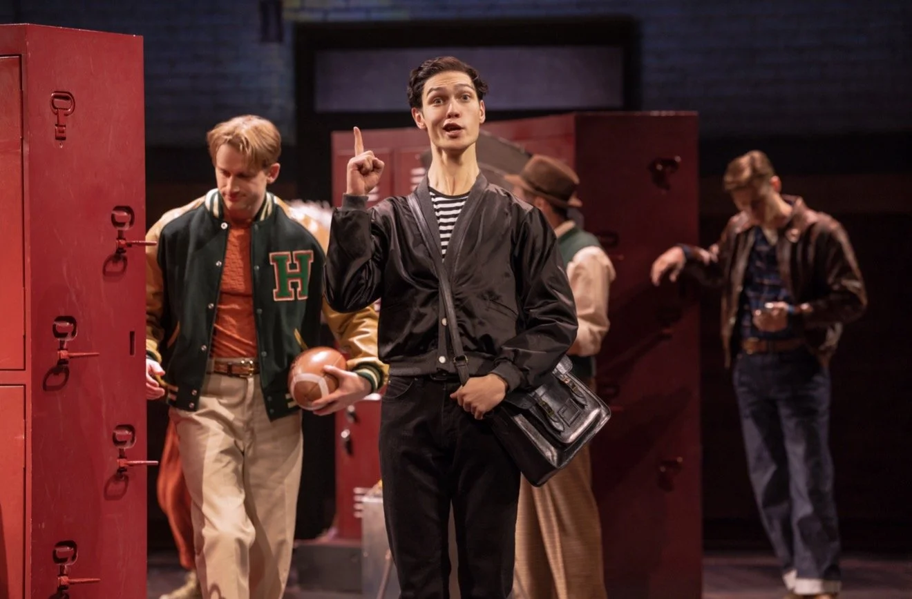 Young man in a black jacket and striped shirt speaking or singing on stage, with four other young men dressed in vintage clothing and varsity jackets, some holding footballs, in a school locker room setting.