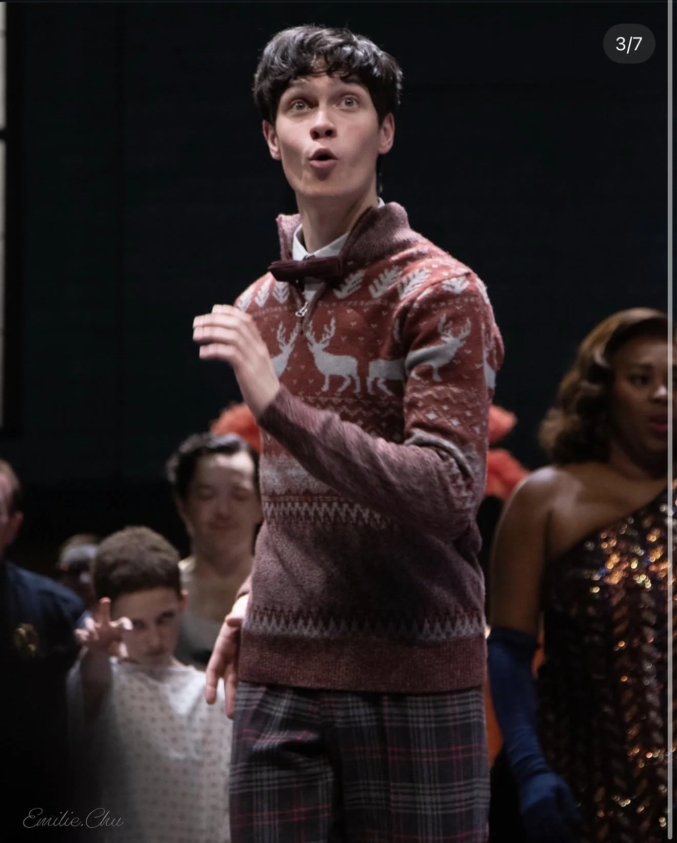 A young man with dark, curly hair and light skin is wearing a festive sweater with reindeer and snowflake patterns and a bow tie, standing among a group of people in a lively setting, possibly at a holiday event.