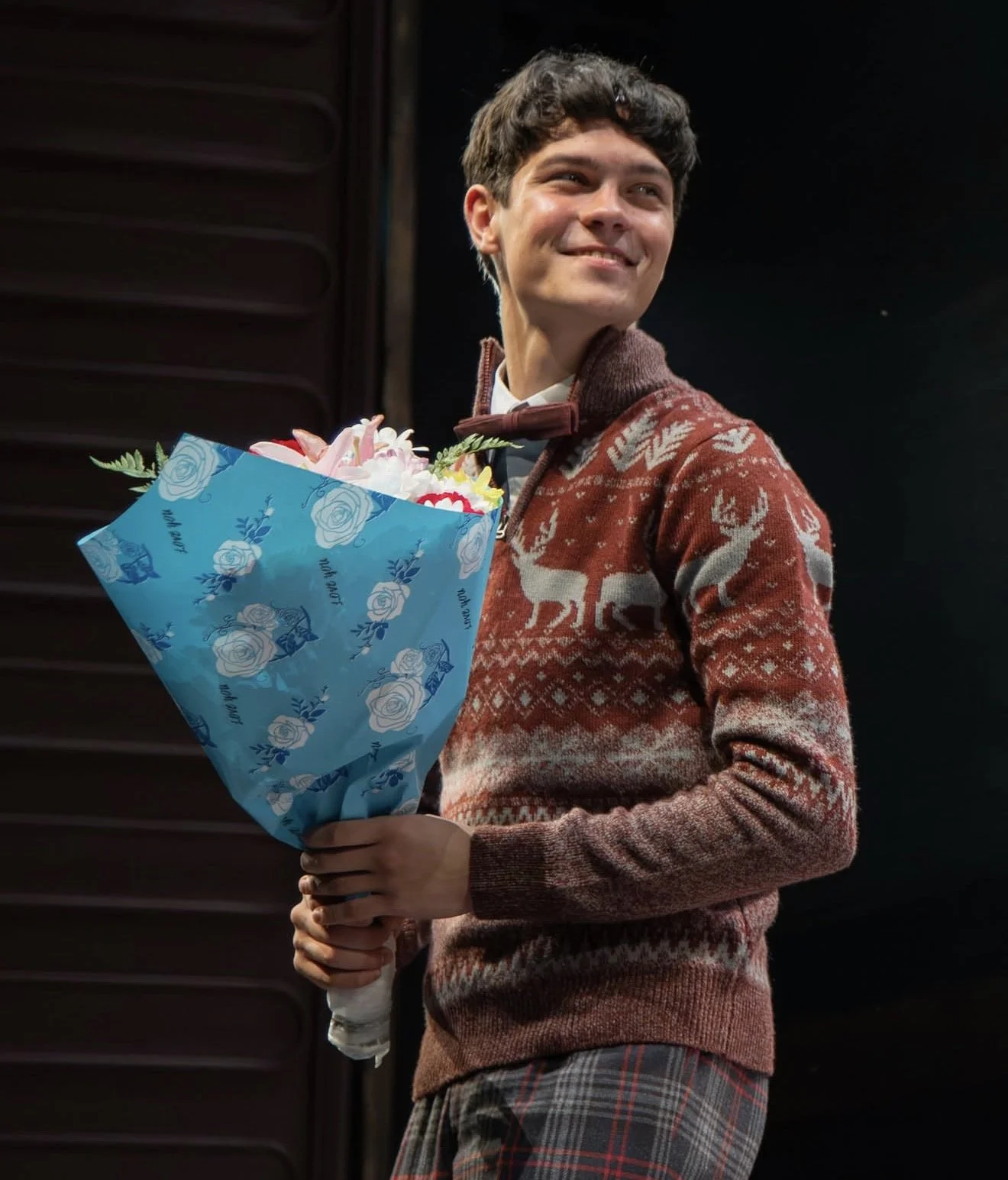 A young man smiling while holding a colorful bouquet of flowers, dressed in a festive sweater with reindeer patterns and plaid pants, standing against a dark background.