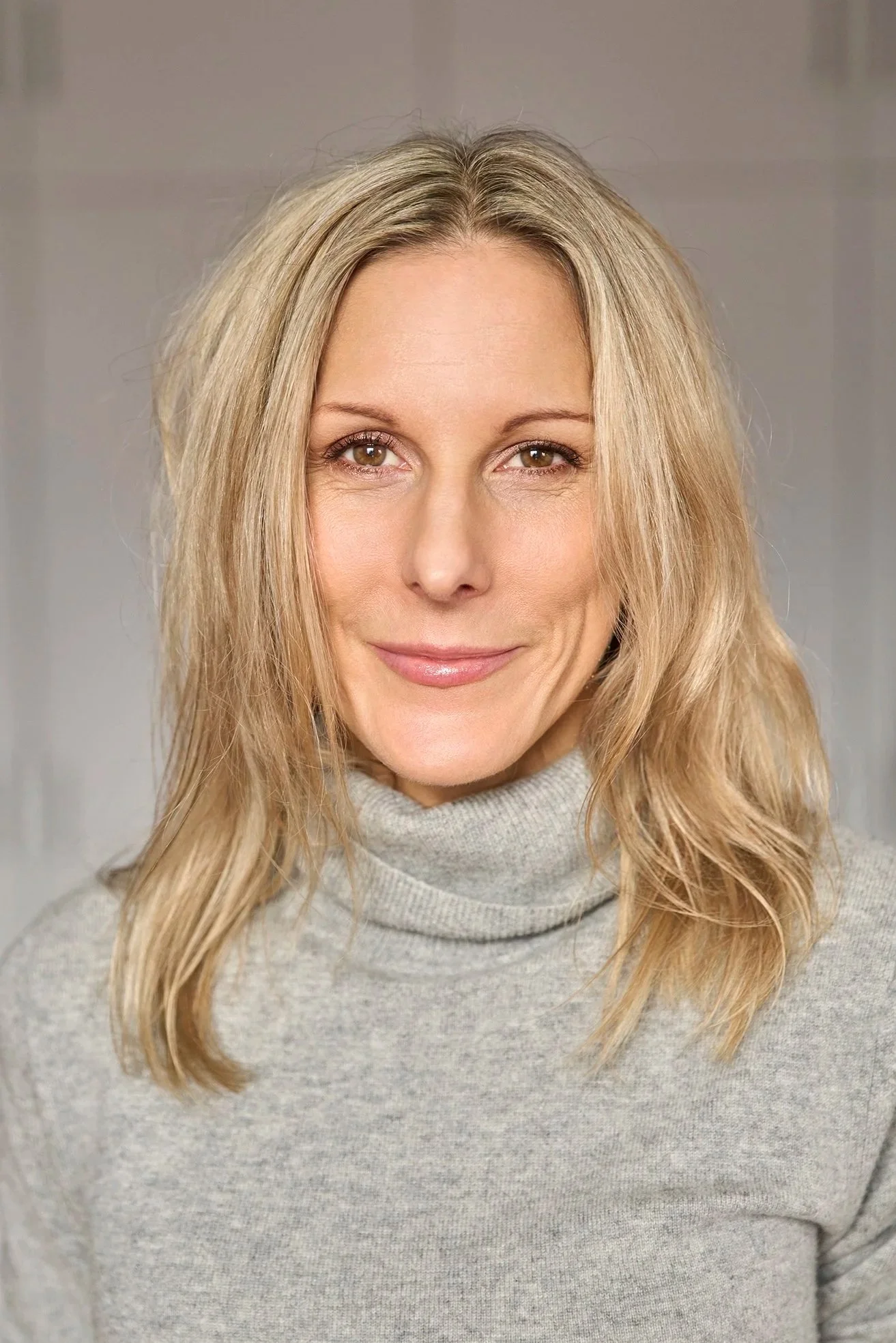 A woman with long blonde hair, light skin, and a warm smile, wearing a light gray turtleneck sweater.