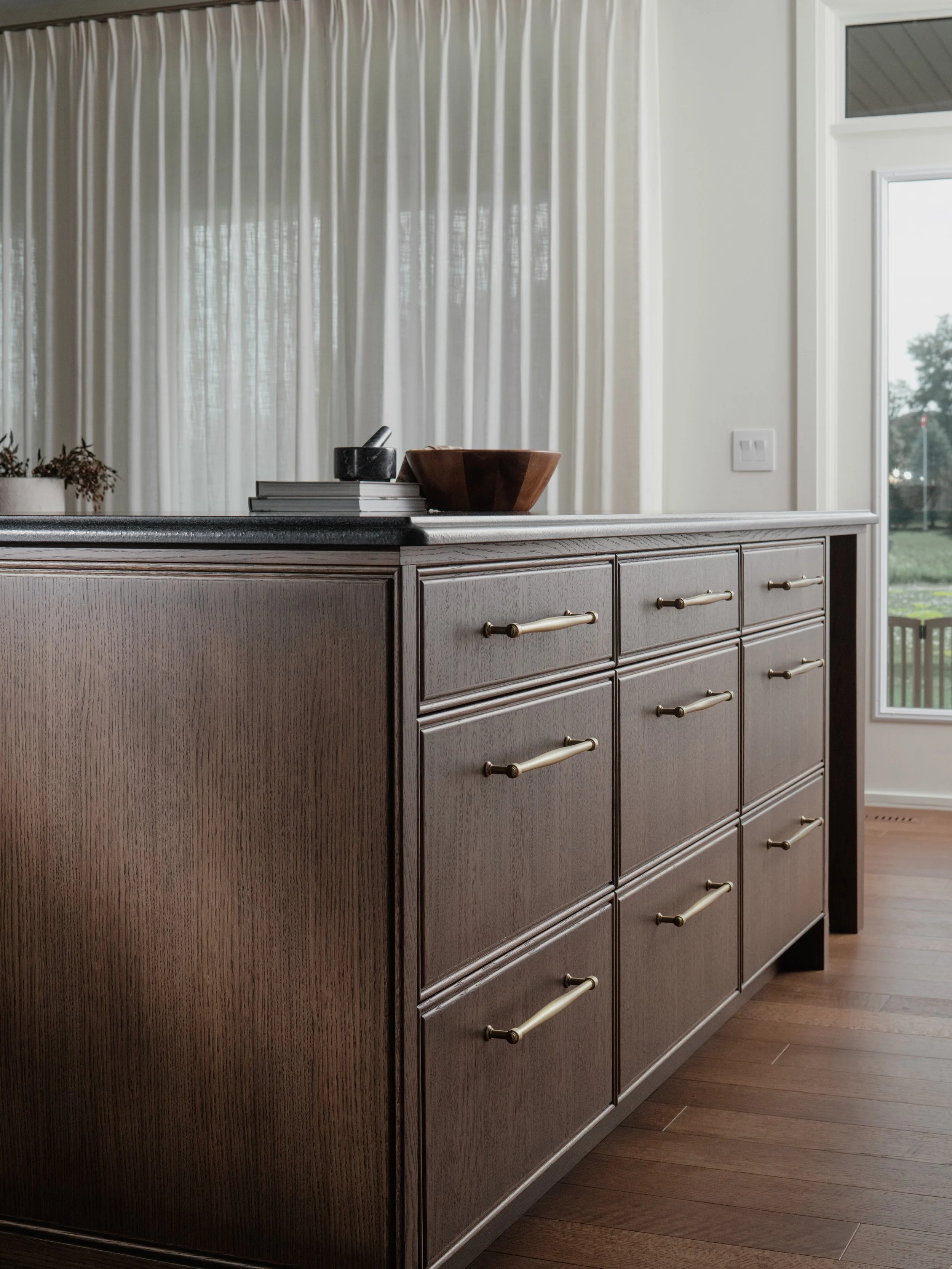 There&rsquo;s something about a dark wood kitchen that feels both bold and timeless. 

For this island, we leaned into rich wood tones, warm brass details and a contrasting dark countertop, balanced with soft creamy cabinetry and drapery. The result 