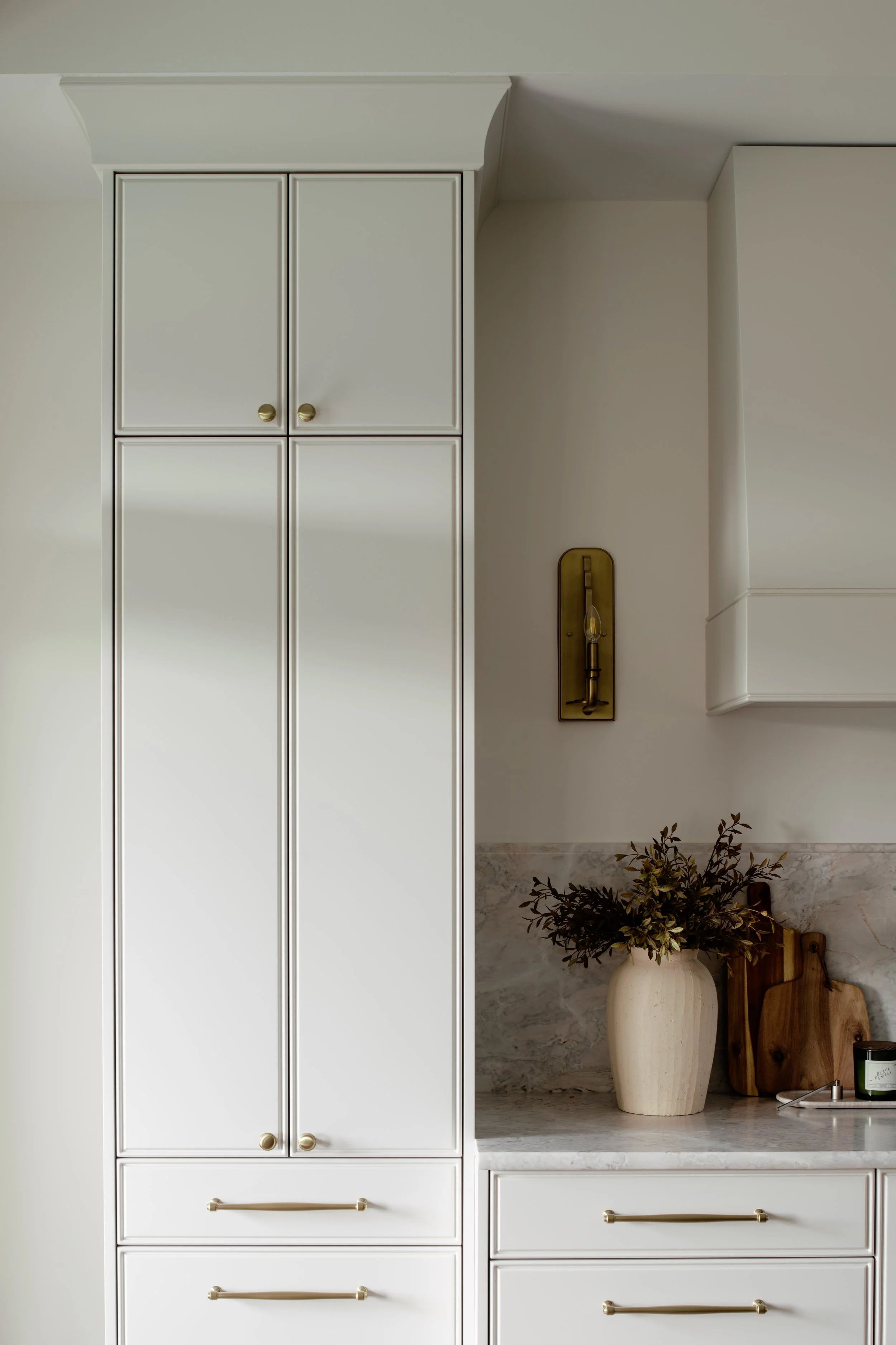 Kitchen ceiling, white cabinets with gold handles, marble countertop, decorative plants, wooden cutting boards, and a vintage-style wall sconce.