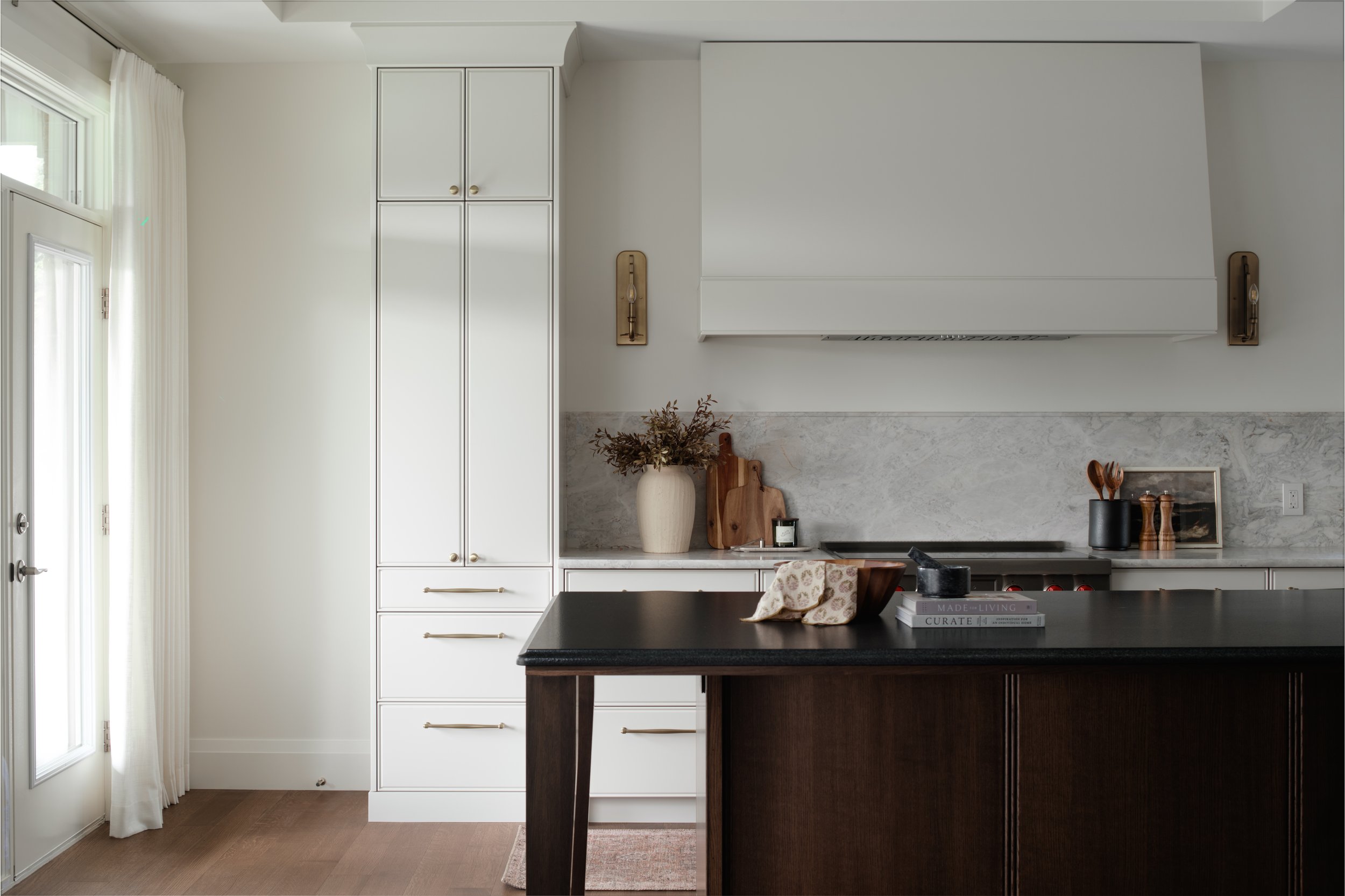 Kitchen with white cabinetry, marble backsplash, black countertop, and kitchen accessories like a vase with dried flowers and books, and a dark wooden island.