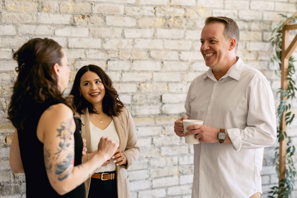 Three people, two women and one man, are standing and talking indoors, smiling and holding mugs, against a white brick wall.