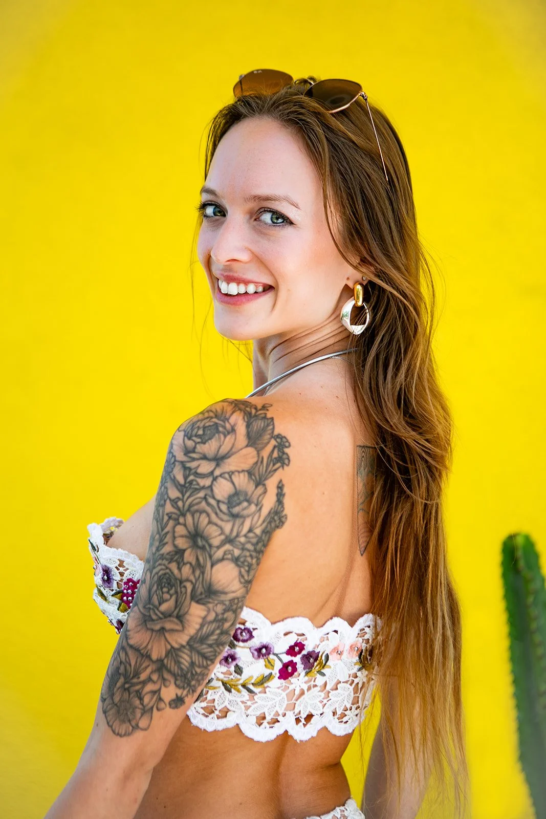 Woman with a floral arm tattoo wearing a lace top, smiling against a yellow background. Indicated CEO of Suhu.