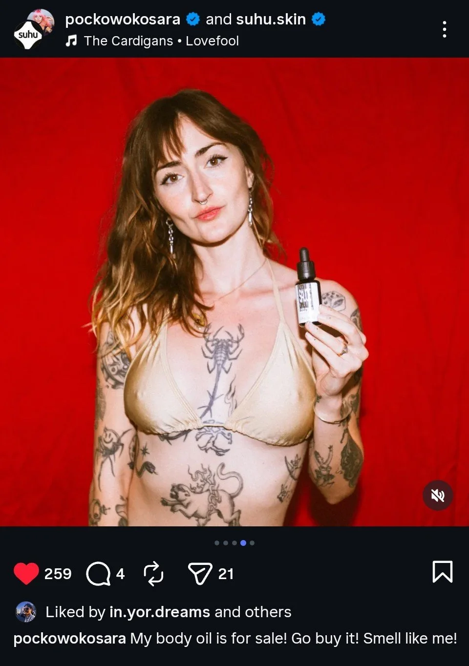 A woman with tattoos holding a small dropper bottle of body oil. She has shoulder-length hair, wears a beige bra, and stands against a red background.