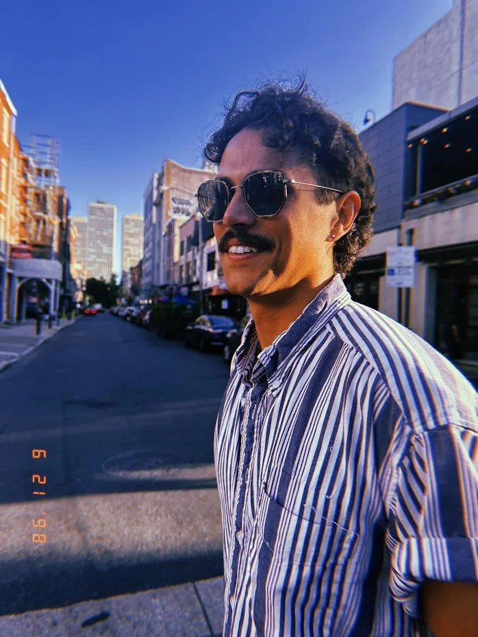 Polaroid style image of a man wearing sunglasses and a striped button-down shirt. The background is a city street.