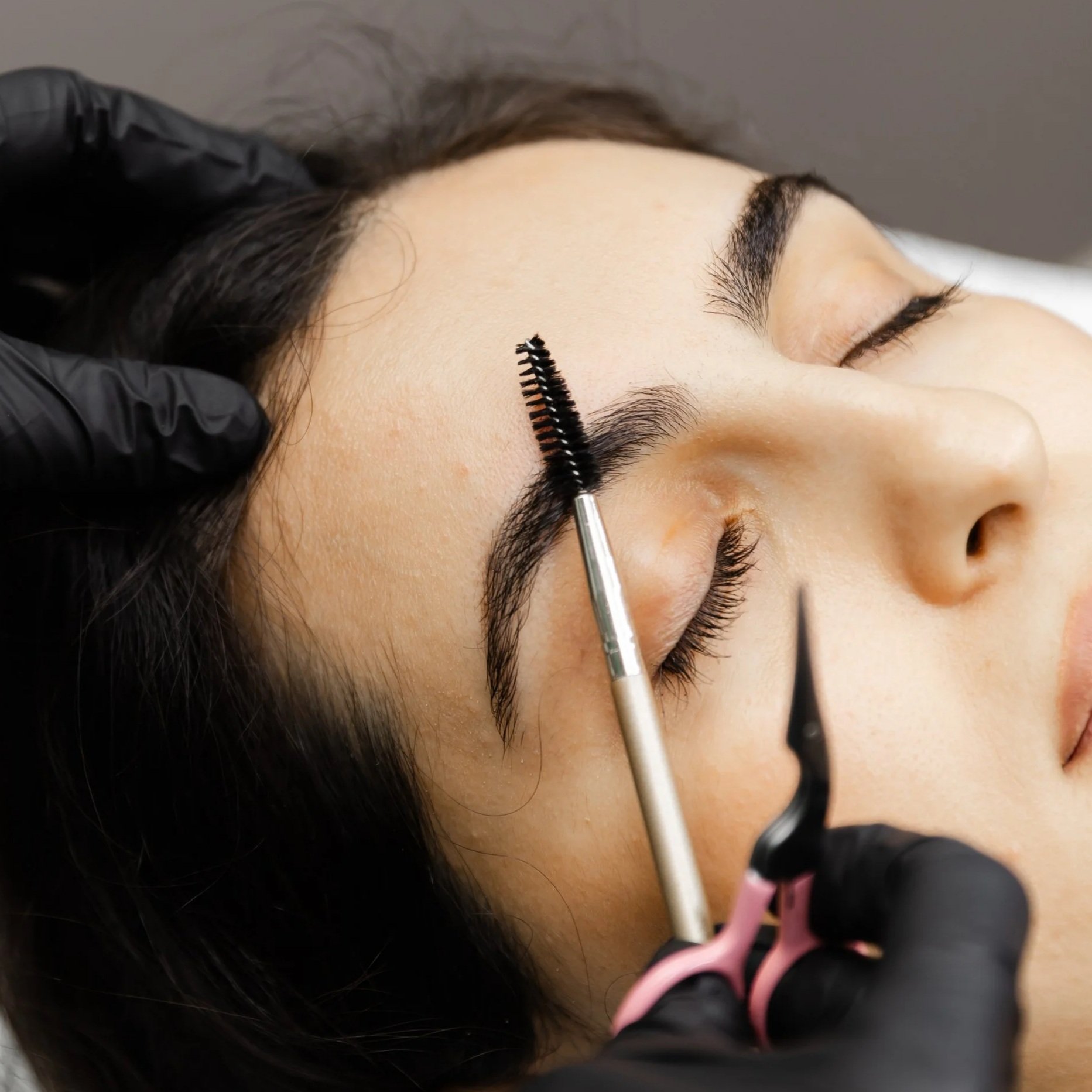 A person lying down with their eyes closed, having their eyebrows shaped with a spoolie brush. The person has dark hair and is wearing black gloves, possibly in a beuaty bar setting.