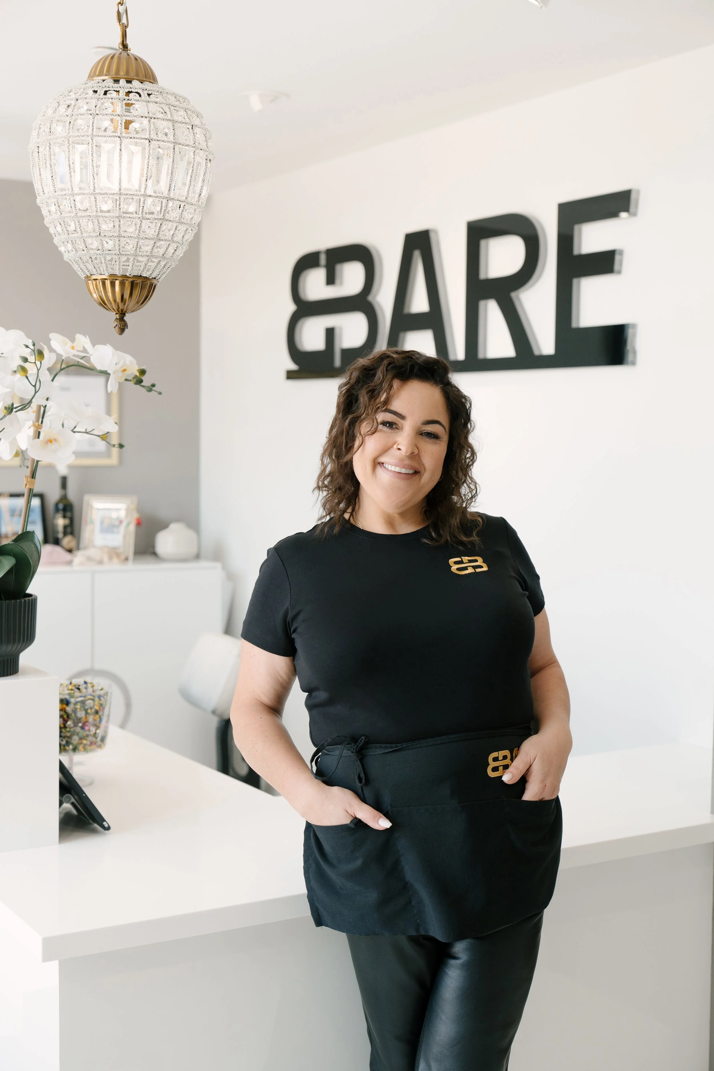 A woman with dark curly hair and a smile, standing in BBare Beauty Bar with a white counter, a large chandelier, orchids, and framed pictures, in front of a wall with a black sign reading 'BBare'.