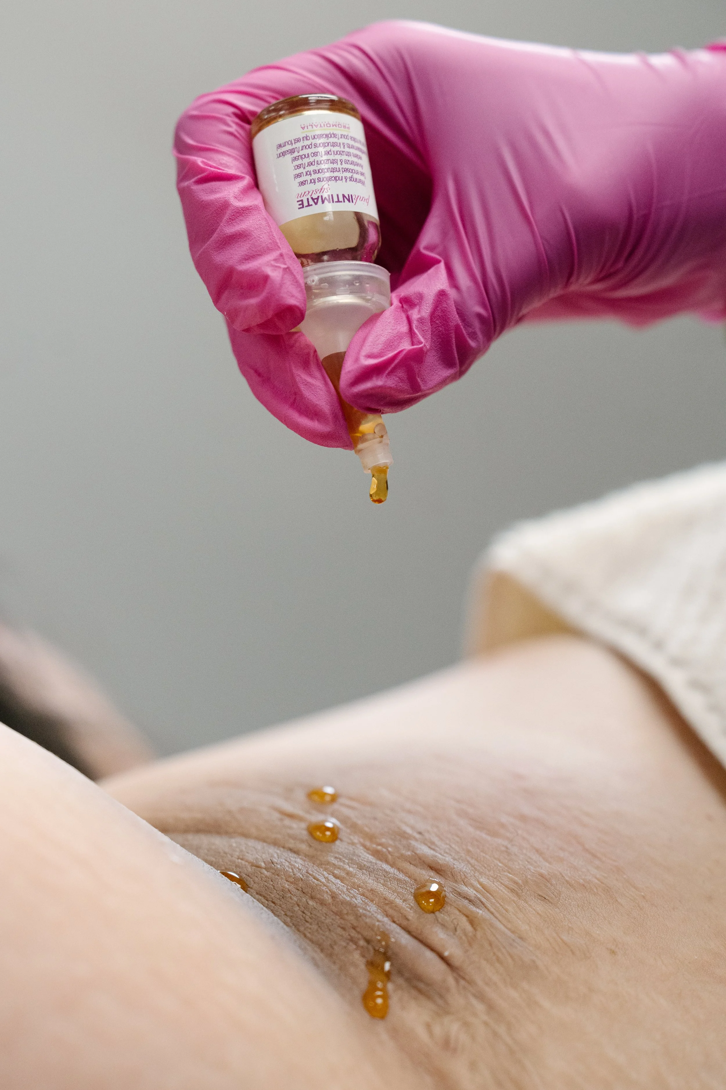 A gloved hand holding a dropper bottle of oil over a person's underarm, dispensing a small amount of oil onto the skin.