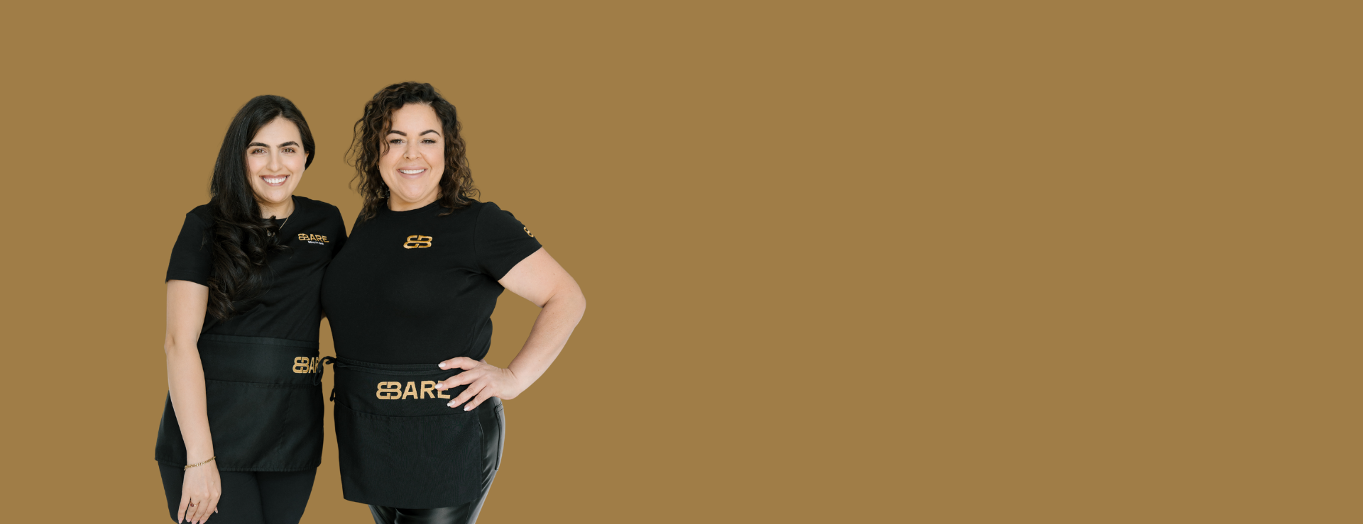 Two women wearing black shirts and aprons with gold logos, standing side by side and smiling against a gold background.