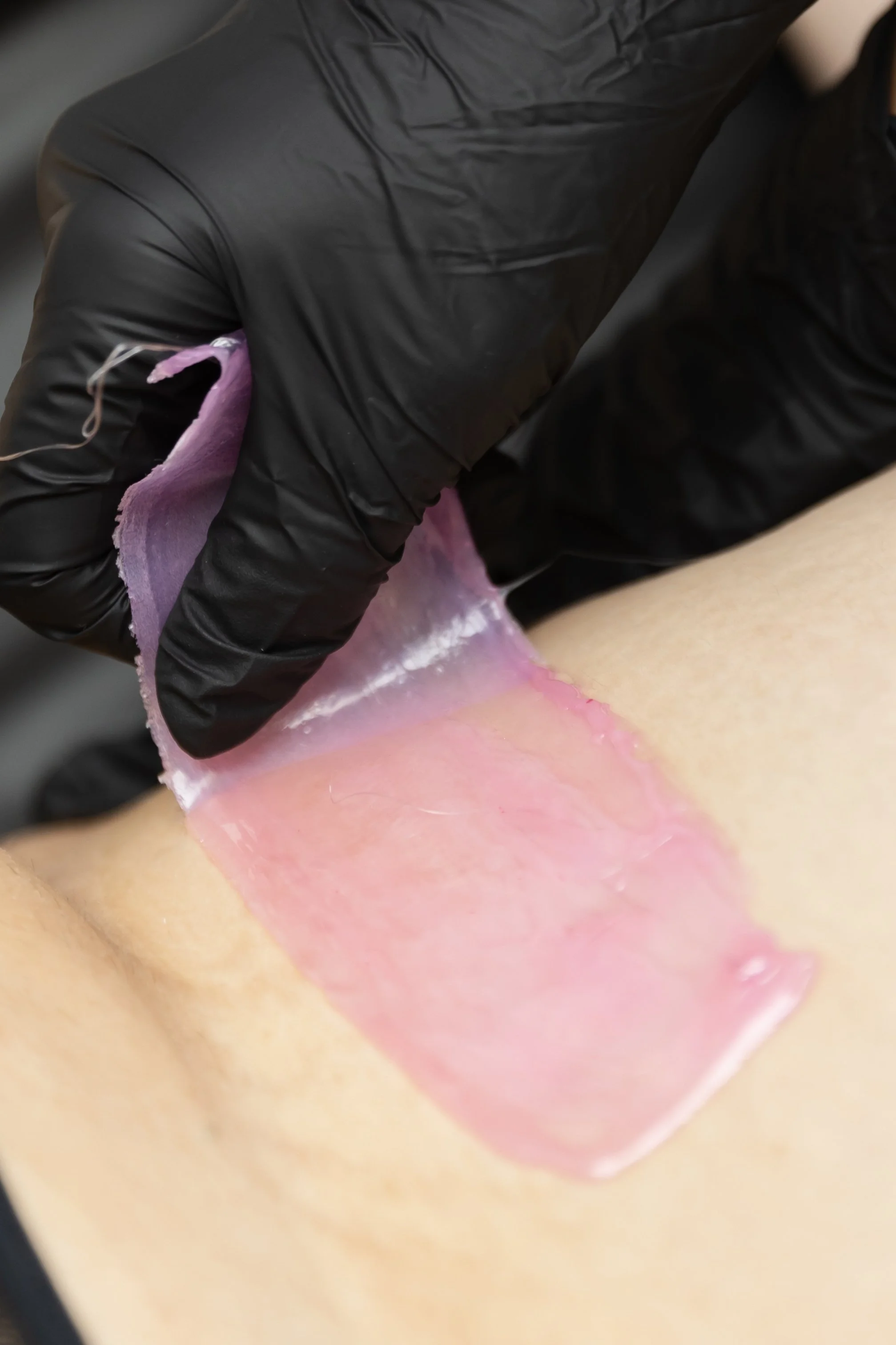 A person receiving a waxing treatment on their leg with a pink wax strip being pulled away.