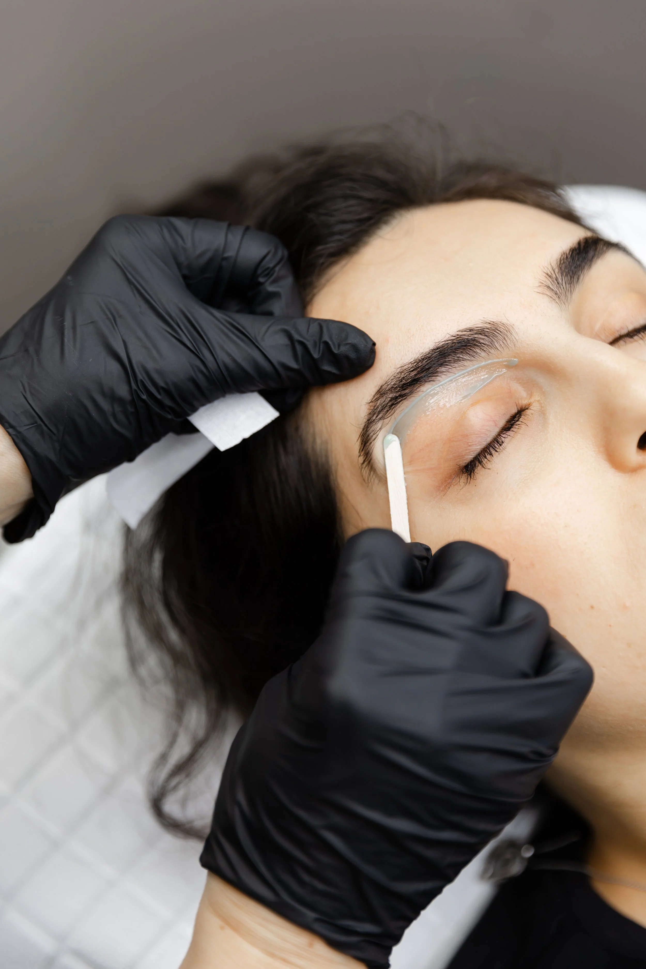 A person is receiving an eyebrow waxing, with a technician applying wax under the client's brows, while the client is lying down with eyes closed.
