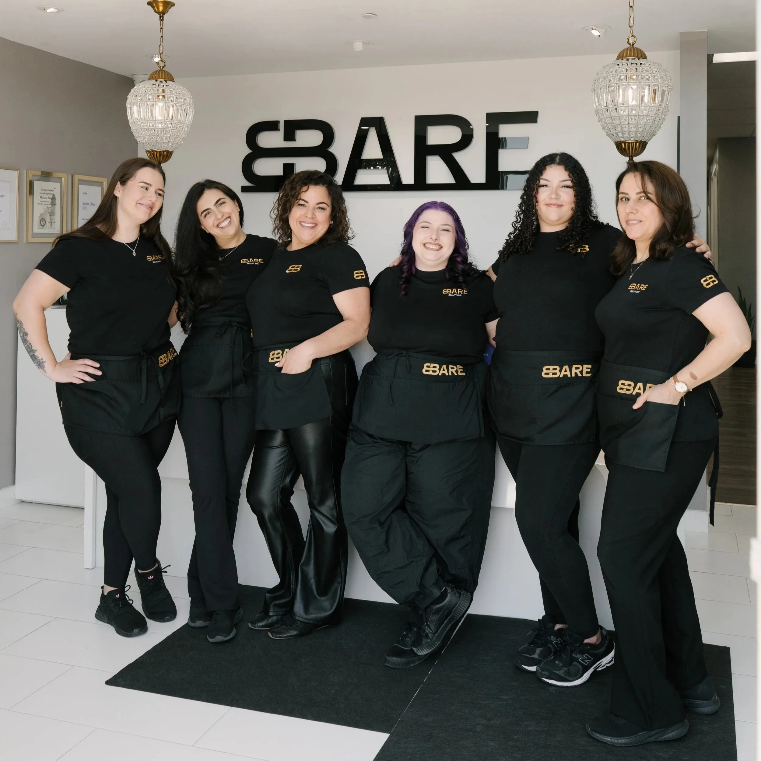 Six women standing together in front of a wall with large black letters spelling 'BARE.' They are smiling, wearing black T-shirts and black pants or leggings. Two women have visible purple hair, and all are wearing athletic shoes. The women are posing casually, some with hands on hips or arms around each other. The setting appears to be indoors, with decorative chandeliers hanging from the ceiling and framed certificates or pictures on the wall to the left.