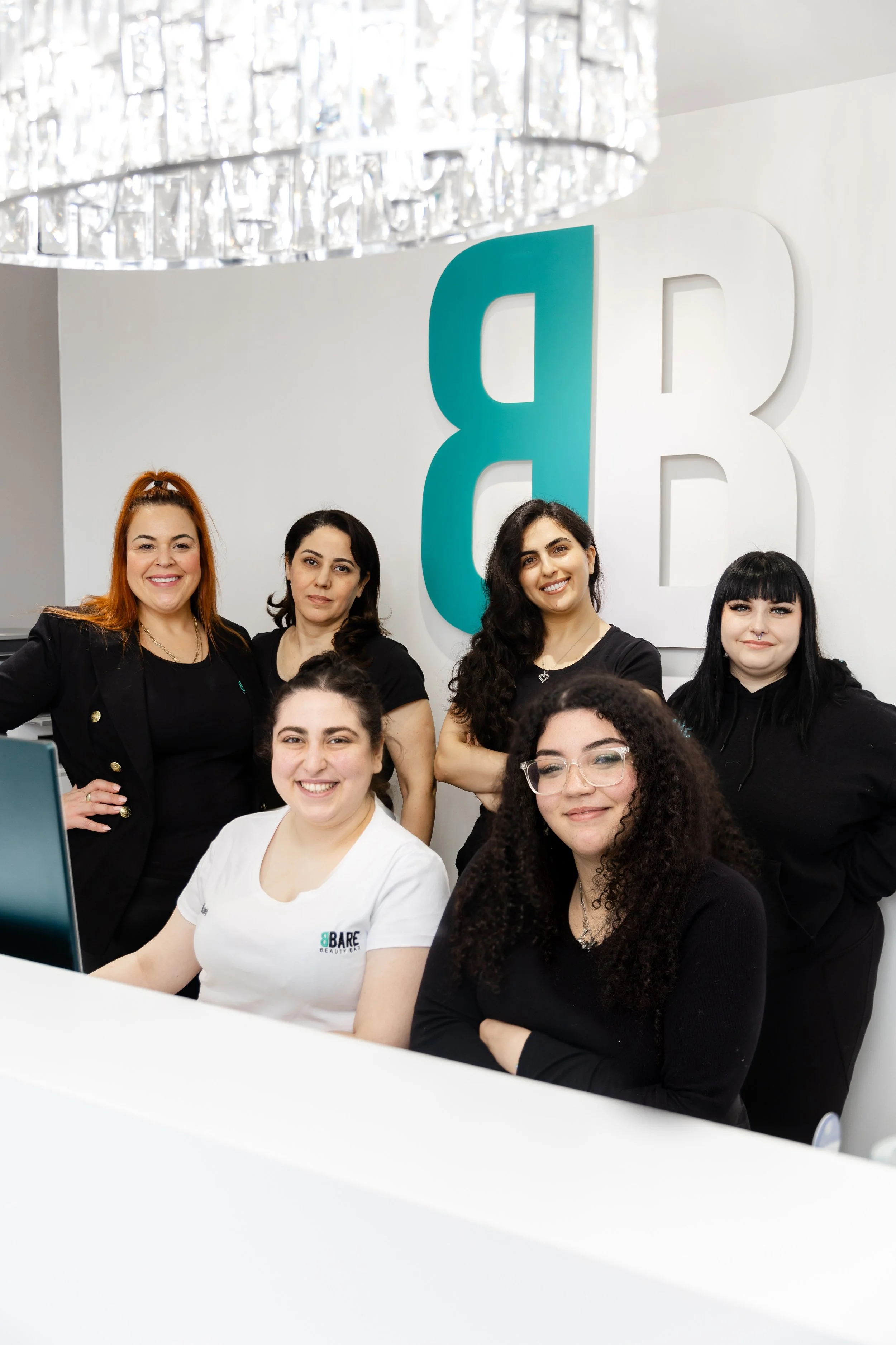 Group of women smiling in an office, with a large blue and white logo of the letter B and B on the wall behind them.