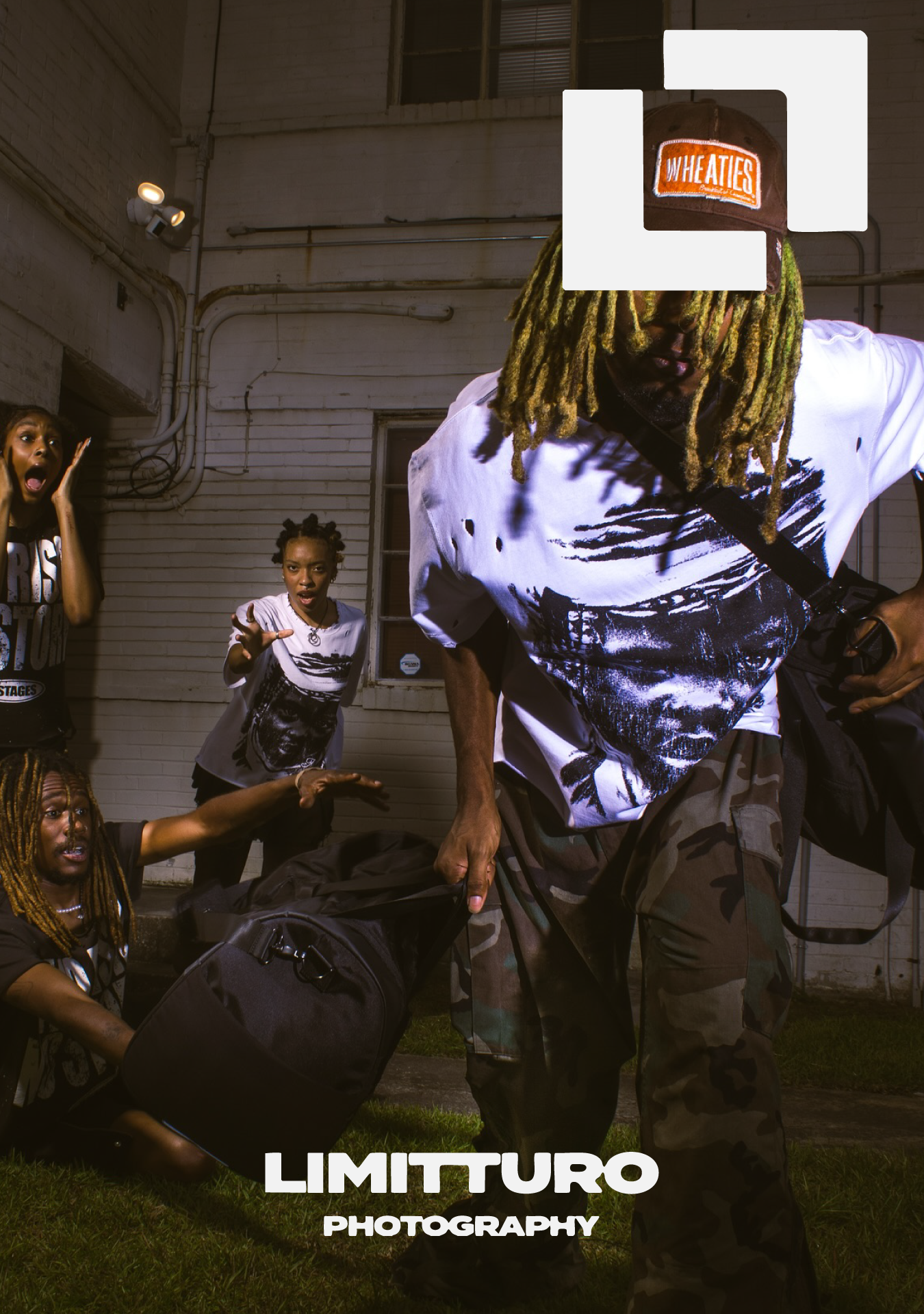 A group of four young people with dreadlocks and casual streetwear posing energetically outdoors at night in front of a white brick wall, with a man in front holding a bag and leaning forward.