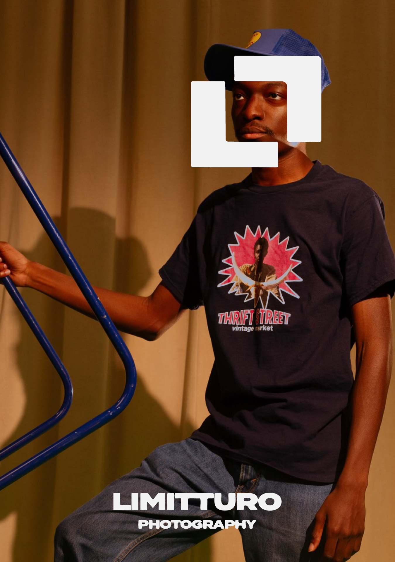 A young man wearing a blue cap, a black T-shirt with a graphic design, and jeans, is standing against a yellow curtain background, holding onto a blue railing.