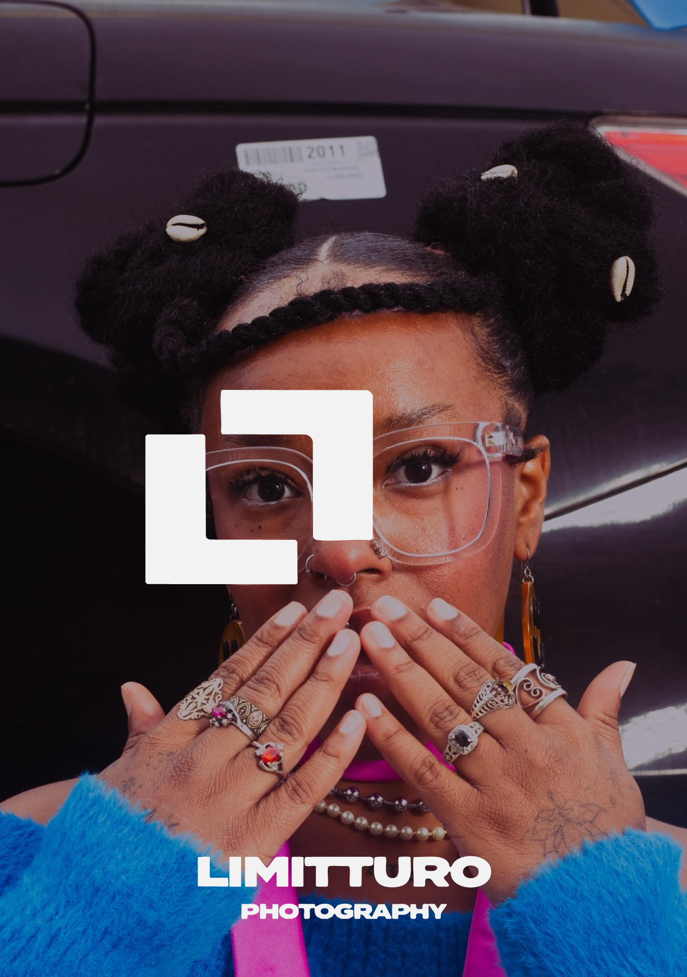 A woman with styled hair featuring hair rollers that have shell-like decorations, wearing glasses, colorful rings, earrings, a pearl necklace, and a fuzzy blue sweater, holding her hands near her face with painted nails, and standing in front of a bl