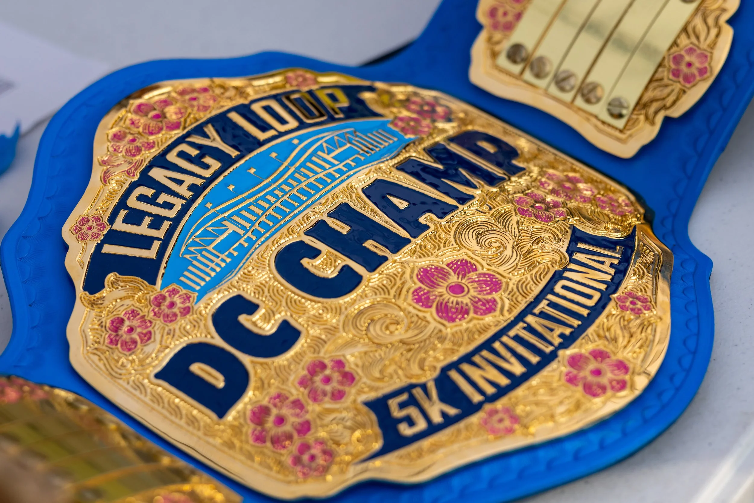 A decorated championship belt with the text 'Legacy Loop DC Champ 5K Invitational' and floral designs.