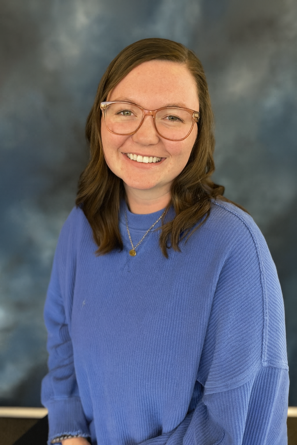 A young woman with brown hair, glasses, and a blue sweater smiling in front of a cloudy sky background.