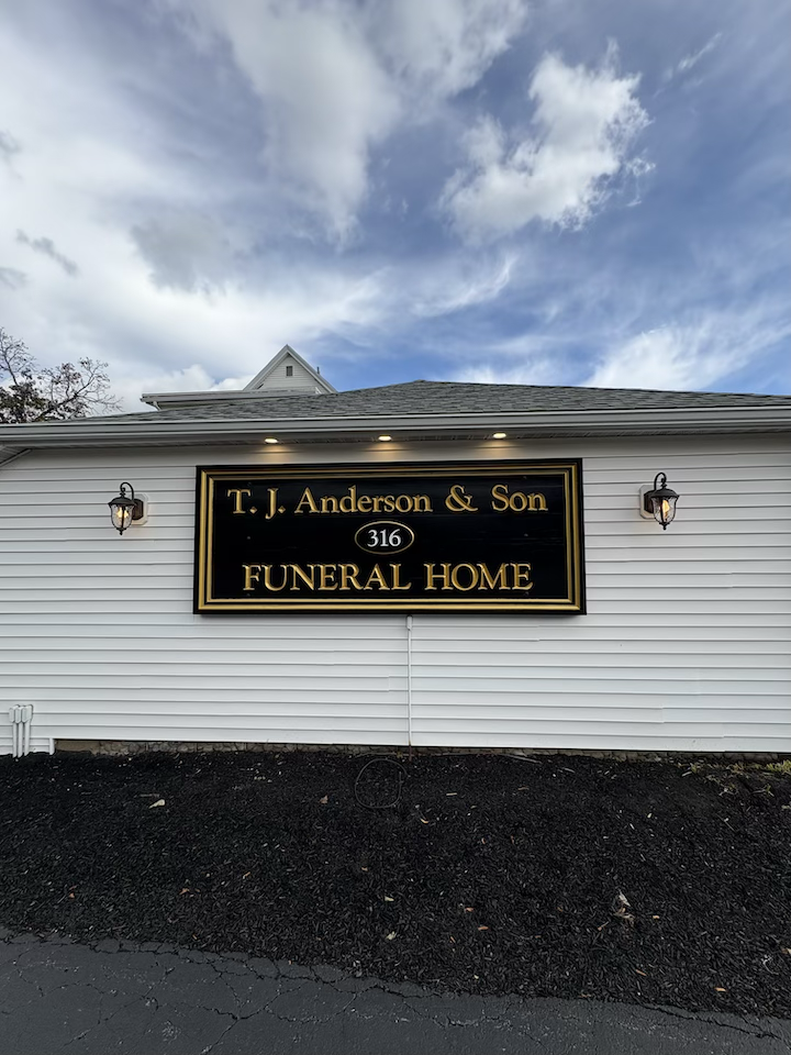 Sign for T. J. Anderson & Son Funeral Home on the exterior of a white building with black trim, under a partly cloudy sky.
