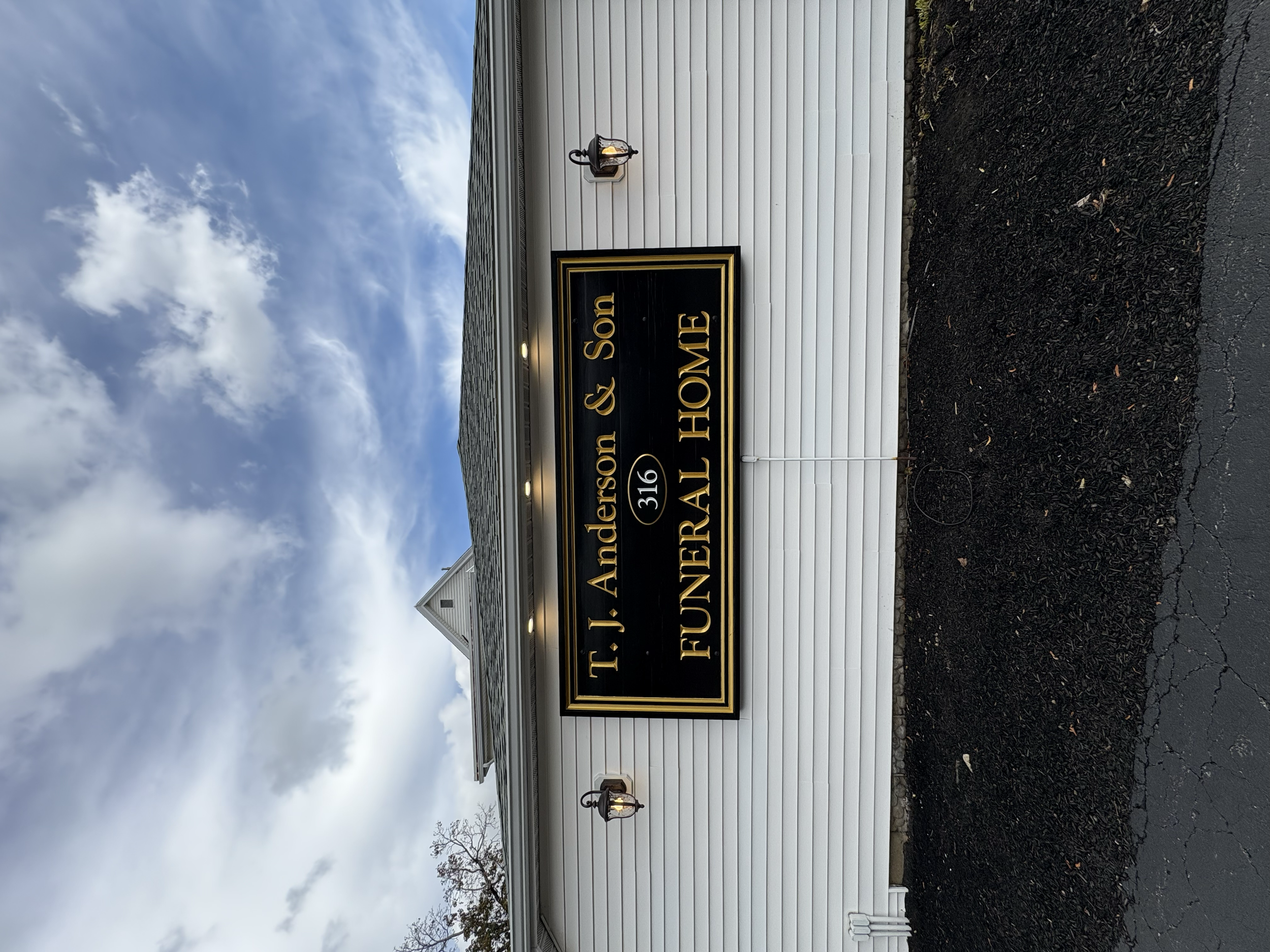 Sign for T.J. Anderson & Son Funeral Home, building with white siding, two outdoor lights, cloudy sky, and asphalt pavement.