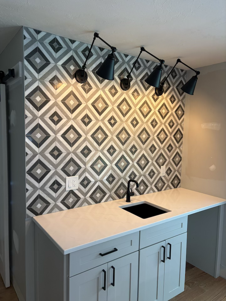 Kitchen area with a geometric black, white, and gray tile backsplash, black adjustable wall-mounted lamps, a white countertop with a black sink, and white cabinets with black handles.
