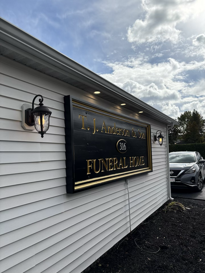 Exterior view of TJ Anderson & Son Funeral Home with a black sign, gold lettering, and two wall-mounted lantern-style lights on either side of the sign, under a partly cloudy sky.